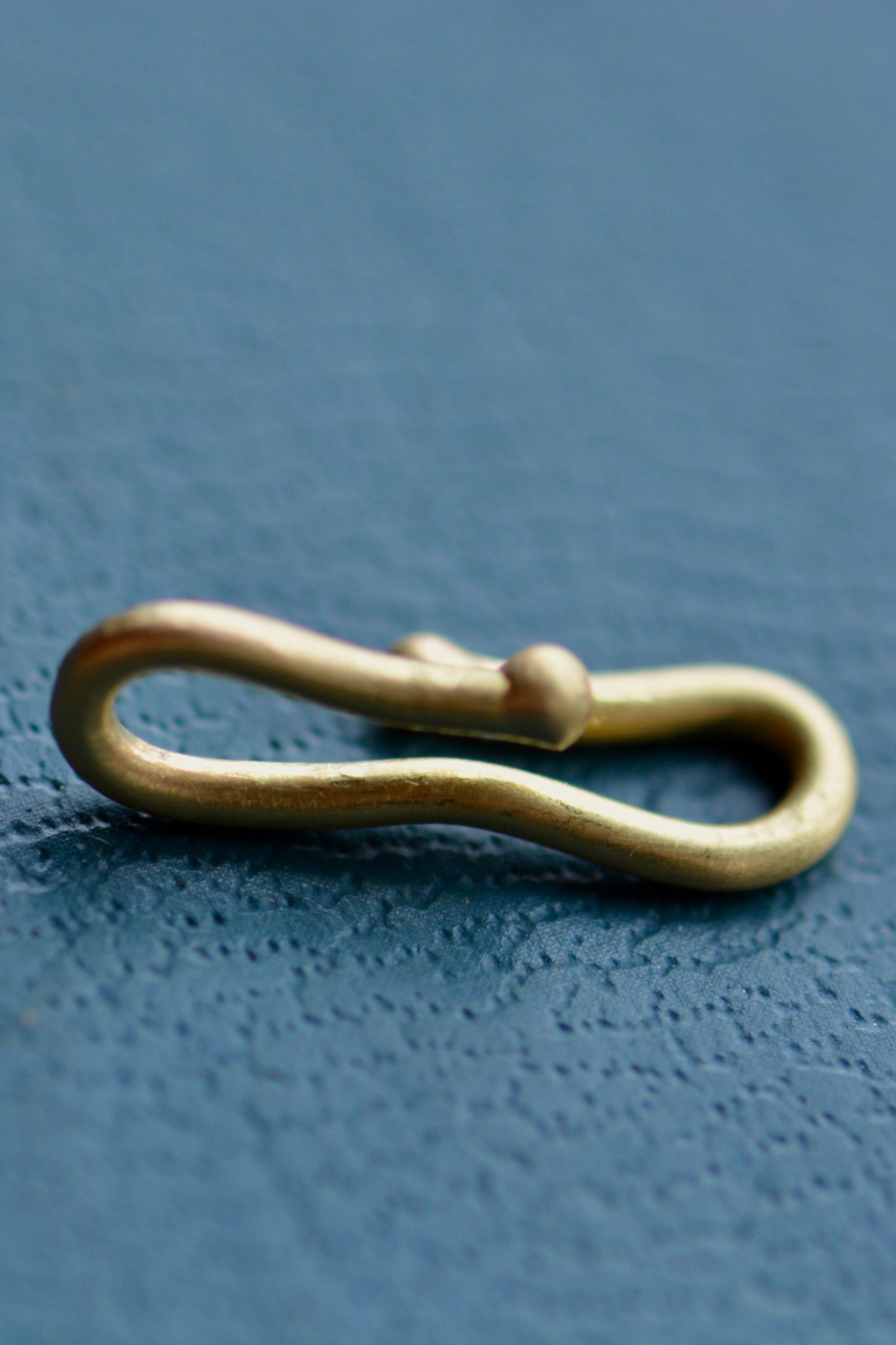 A gold charm connector hook sits on a teal blue textured backdrop.