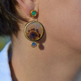 A close-up of a person's ear wearing a gold and oxidized silver dangle earring with a circular natural dendritic agate stone and two small blue and green opals.