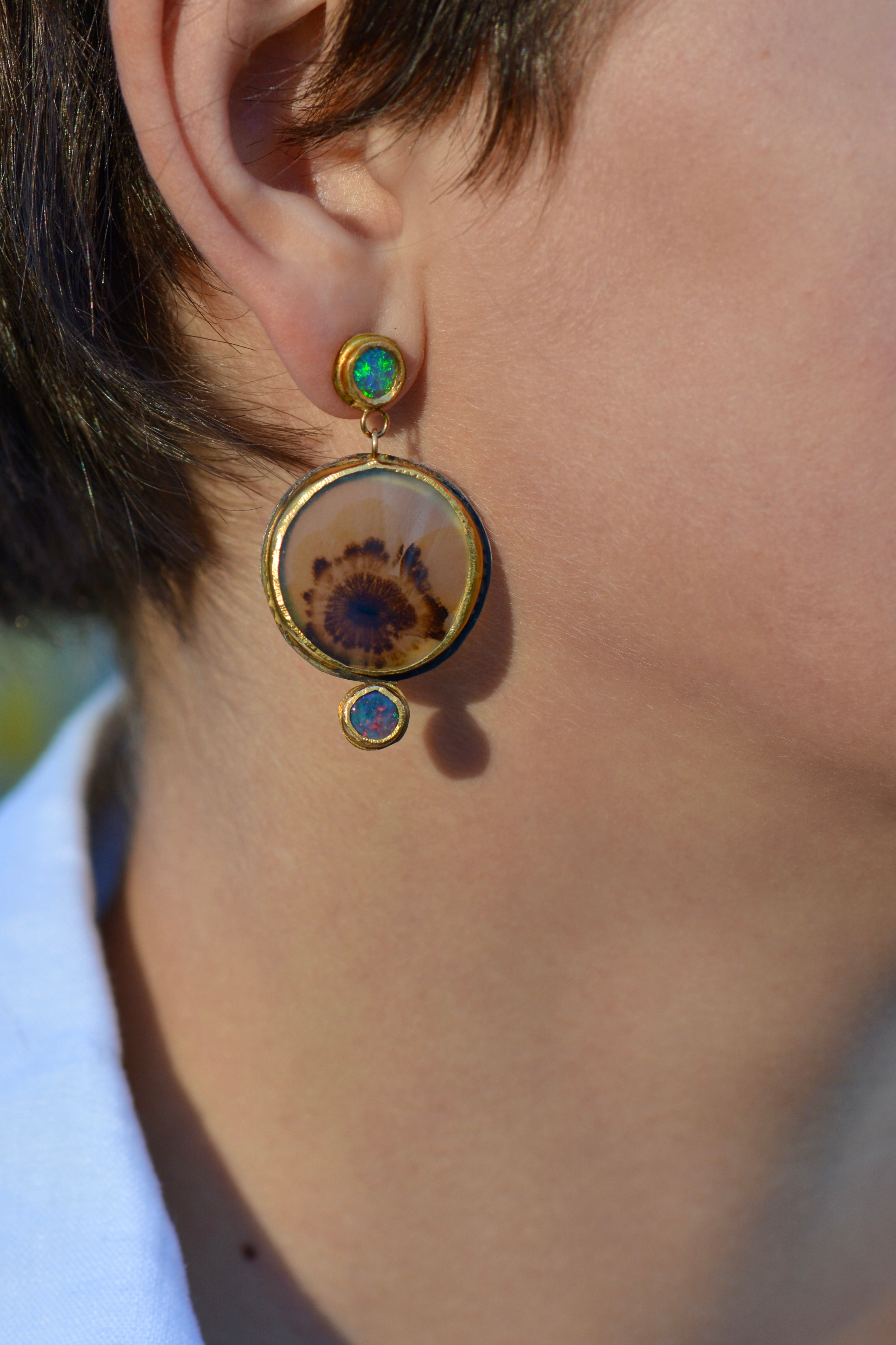 A close-up of a person's ear wearing a gold and oxidized silver dangle earring with a circular natural dendritic agate stone and two small blue and green opals.