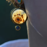 A close-up lifestyle shot of a person's ear wearing a unique gold-toned dandelion dendritic agate pendant earring with small blue opal stones.