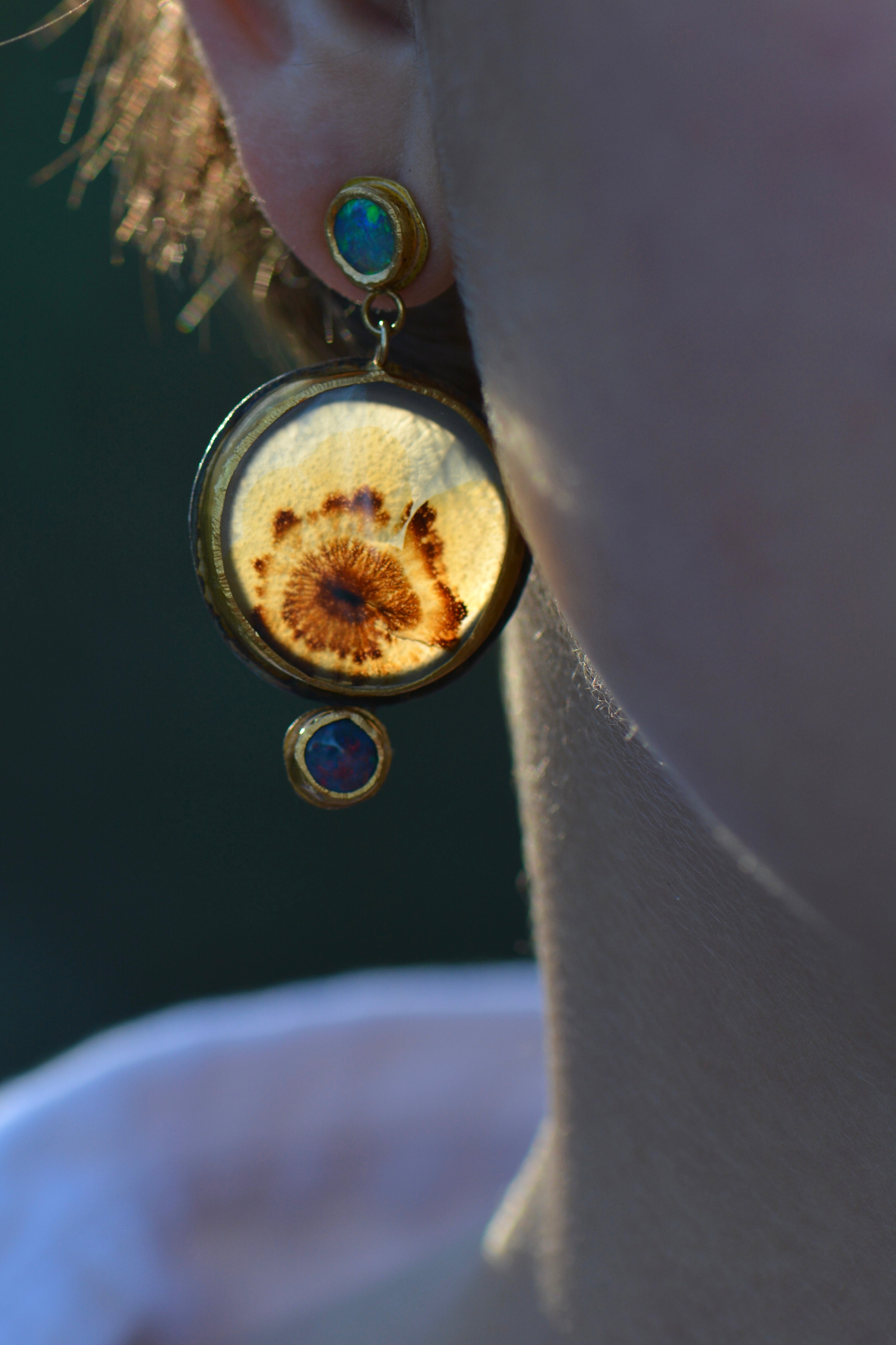 A close-up lifestyle shot of a person's ear wearing a unique gold-toned dandelion dendritic agate pendant earring with small blue opal stones.