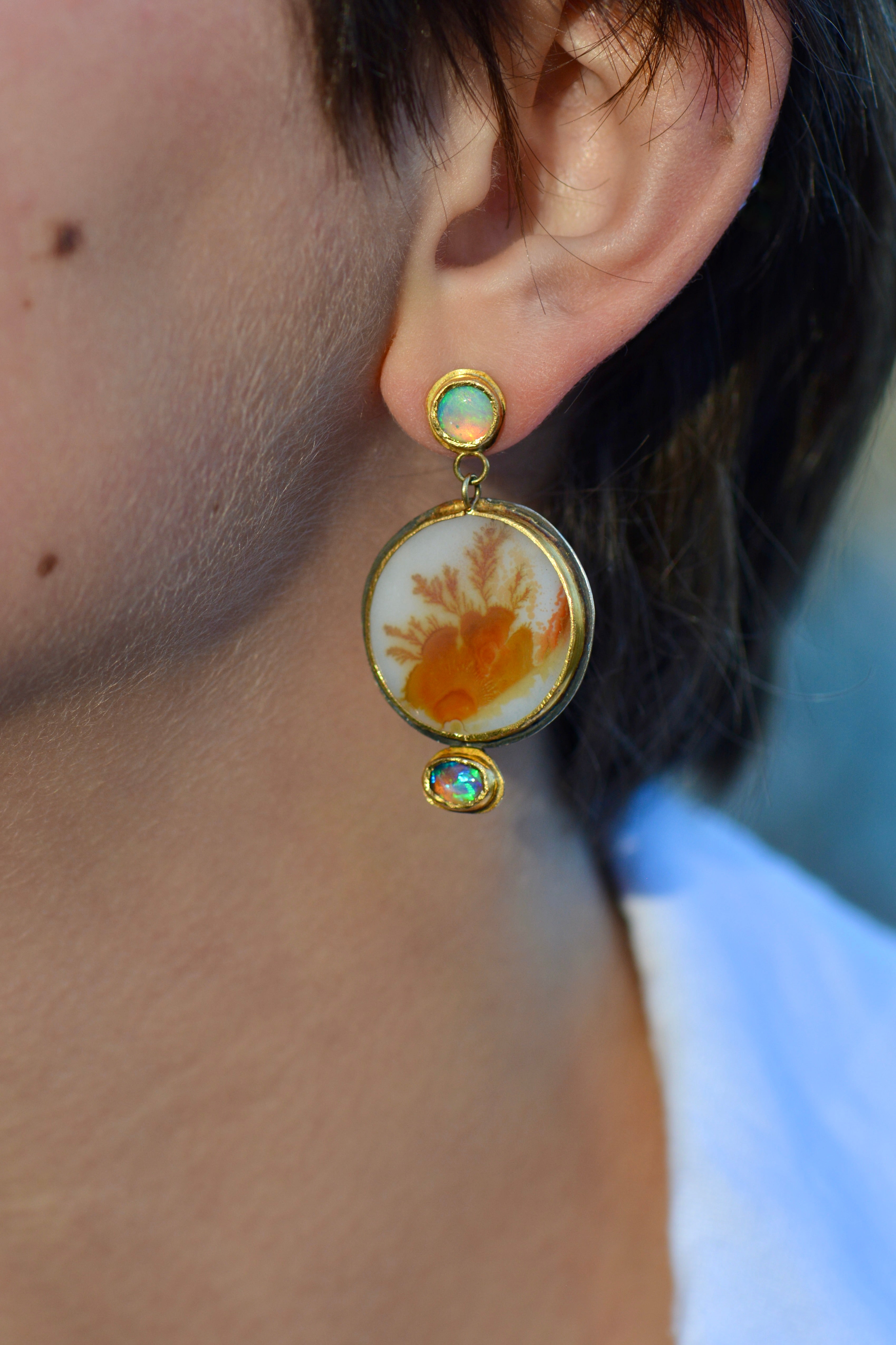 A close-up of a person's ear wearing a dangling earring. The earring has a gold and silver mixed metal setting with a circular yellow-orange floral or coral-like inclusion in a dendritic agate stone, plus small iridescent opal stones at the top and bottom.