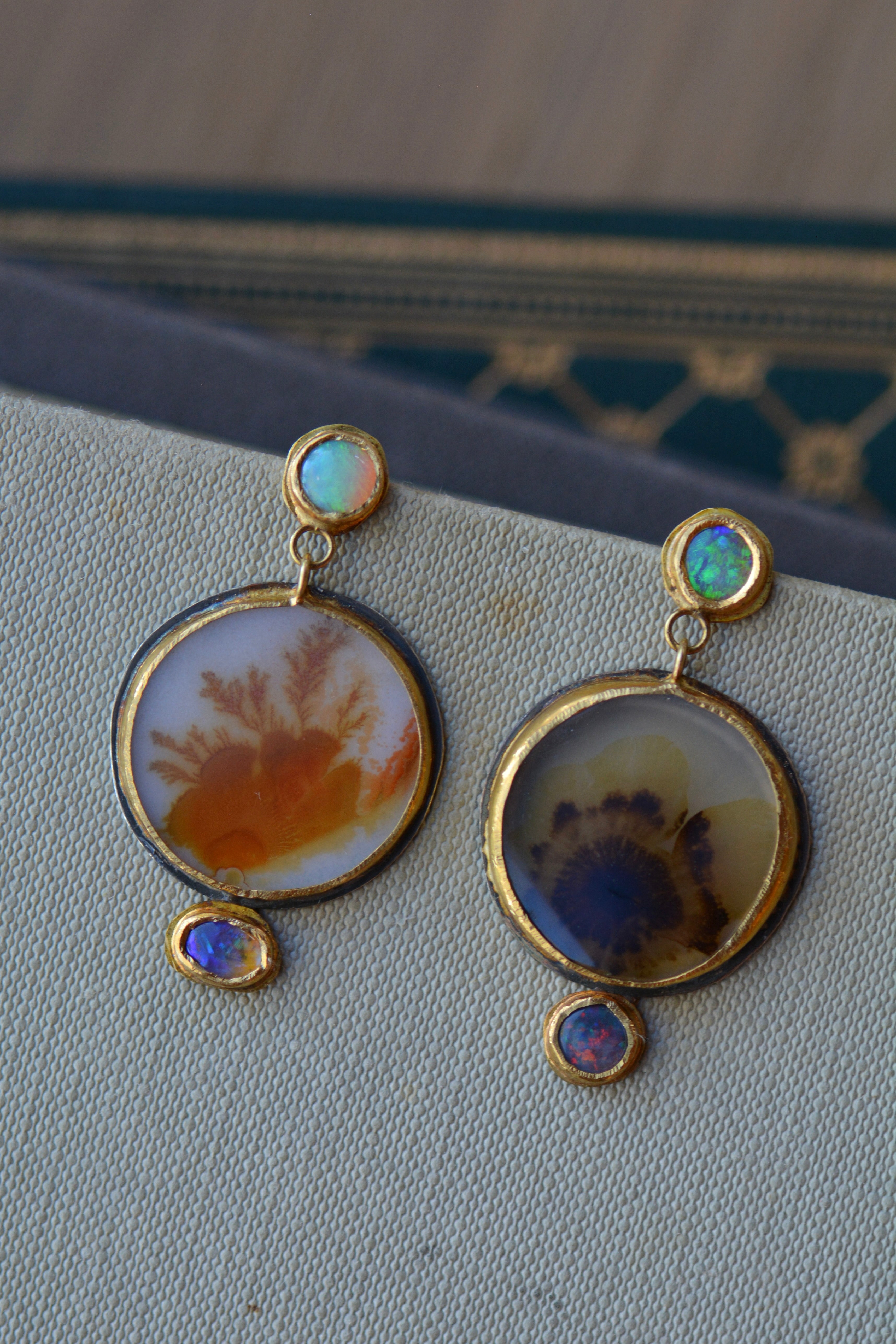 A pair of gold and silver mismatched dangle earrings featuring round agate pendants with natural inclusions, small round opal stones at the top, and oval blue opal stones at the bottom, displayed on a textured light grey surface.