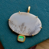 A white agate stone with delicate grey and brown branching patterns in a gold setting with a green and blue accent stone sits on a textured teal backdrop.