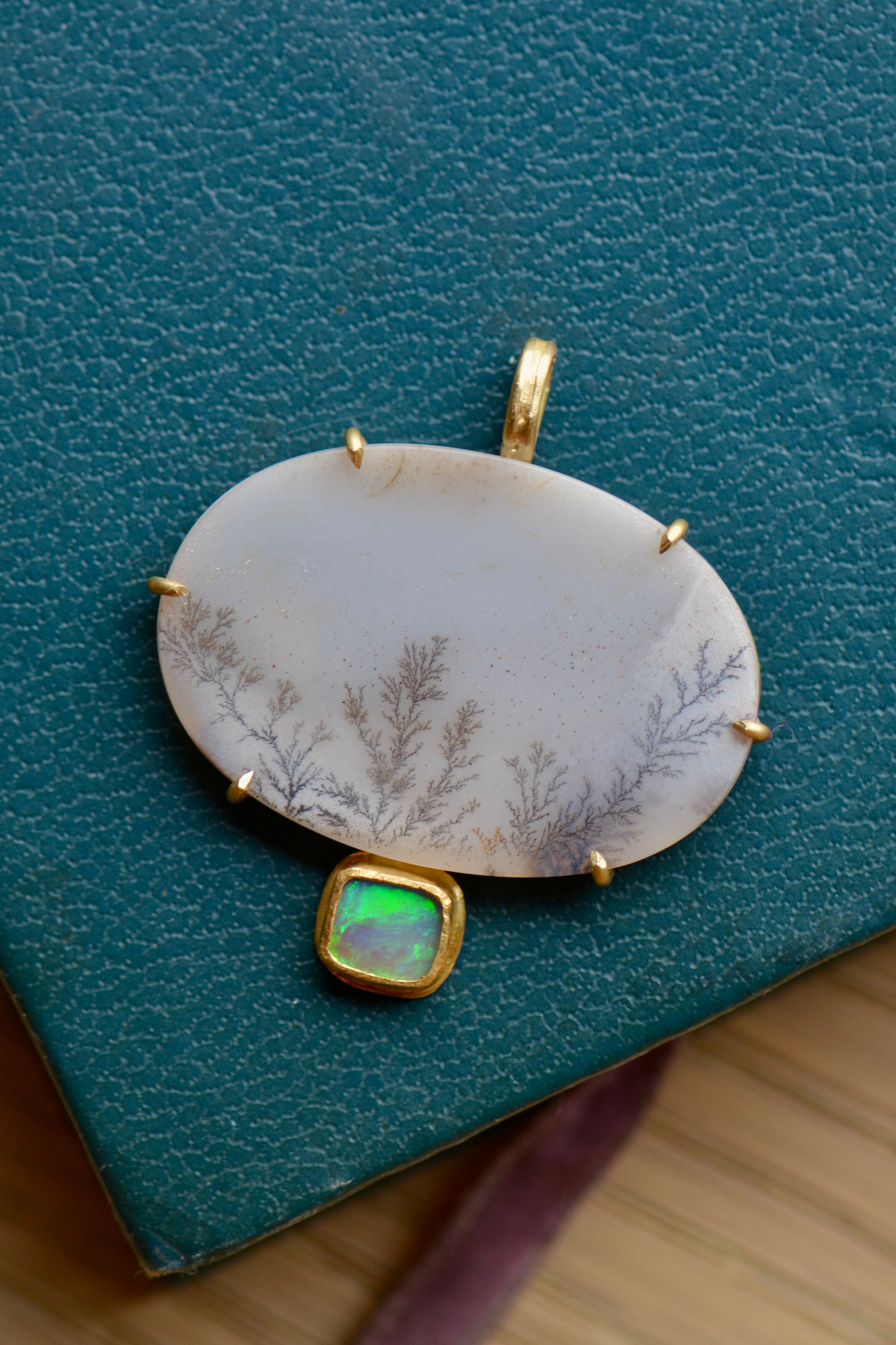 A white agate stone with delicate grey and brown branching patterns in a gold setting with a green and blue accent stone sits on a textured teal backdrop.