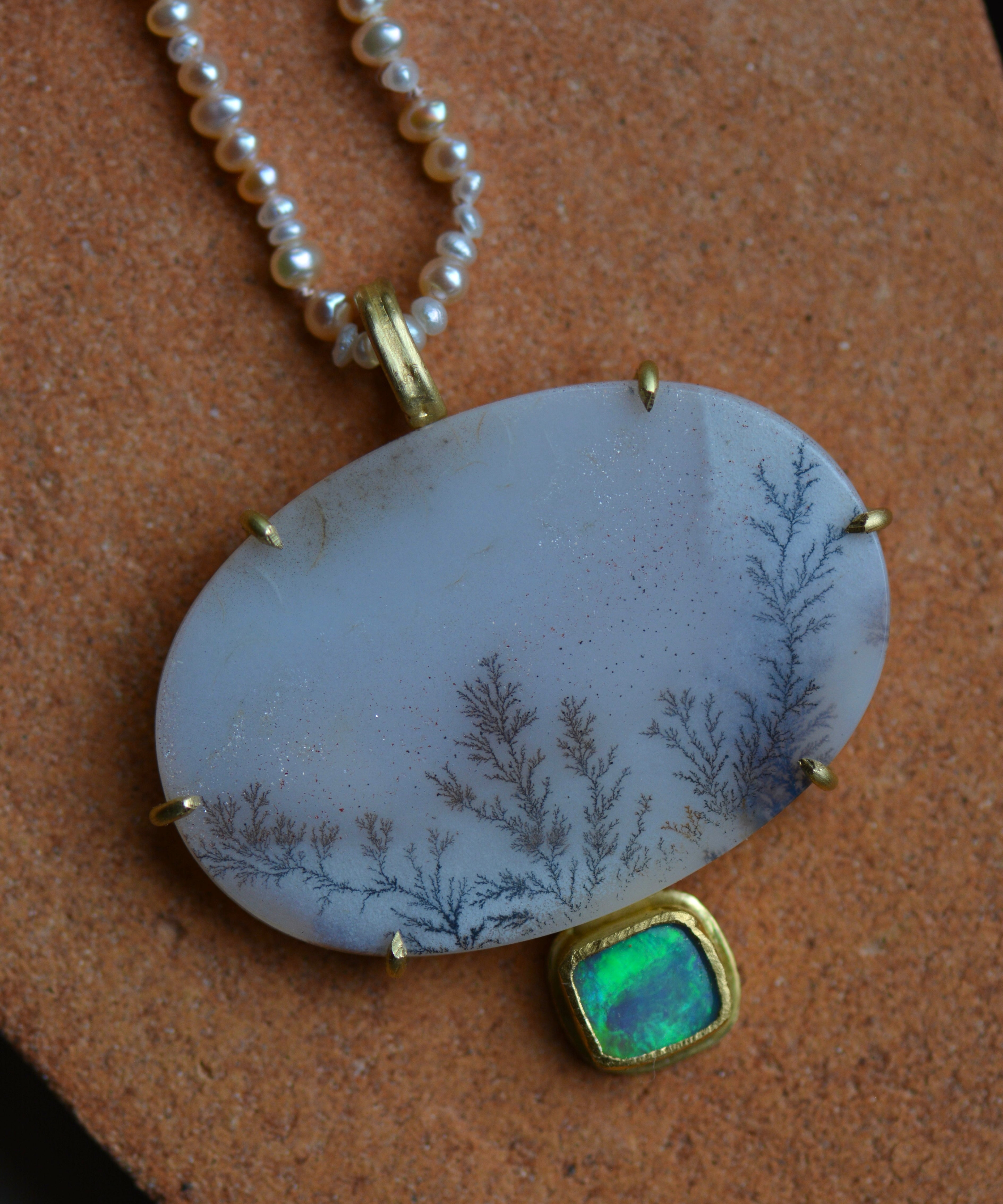 A sparkling dendritic agate oval stone with a blue and green flashing opal stone on the bottom, shown on a pearl beaded strand.