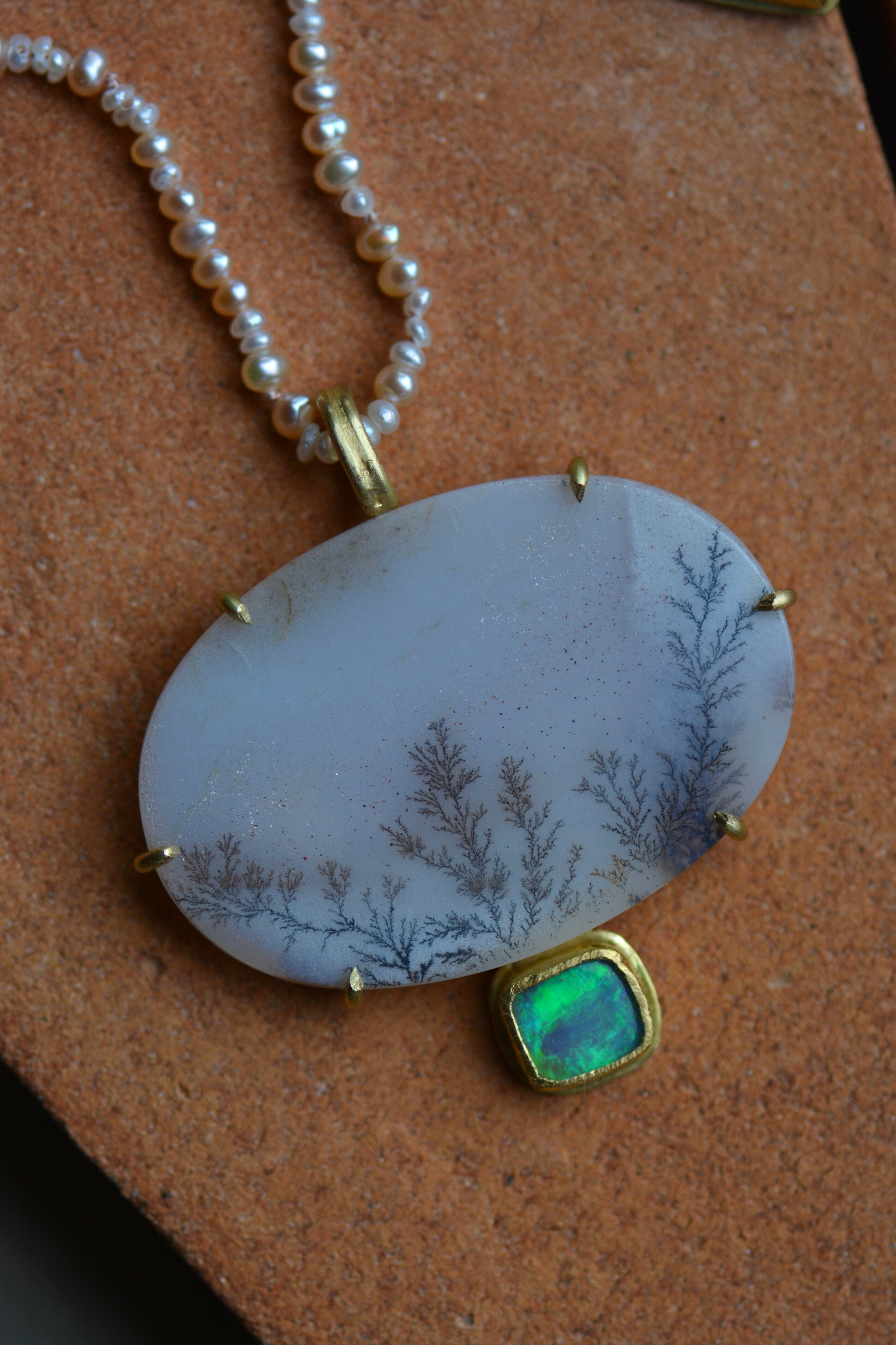 A sparkling dendritic agate oval stone with a blue and green flashing opal stone on the bottom, shown on a pearl beaded strand.