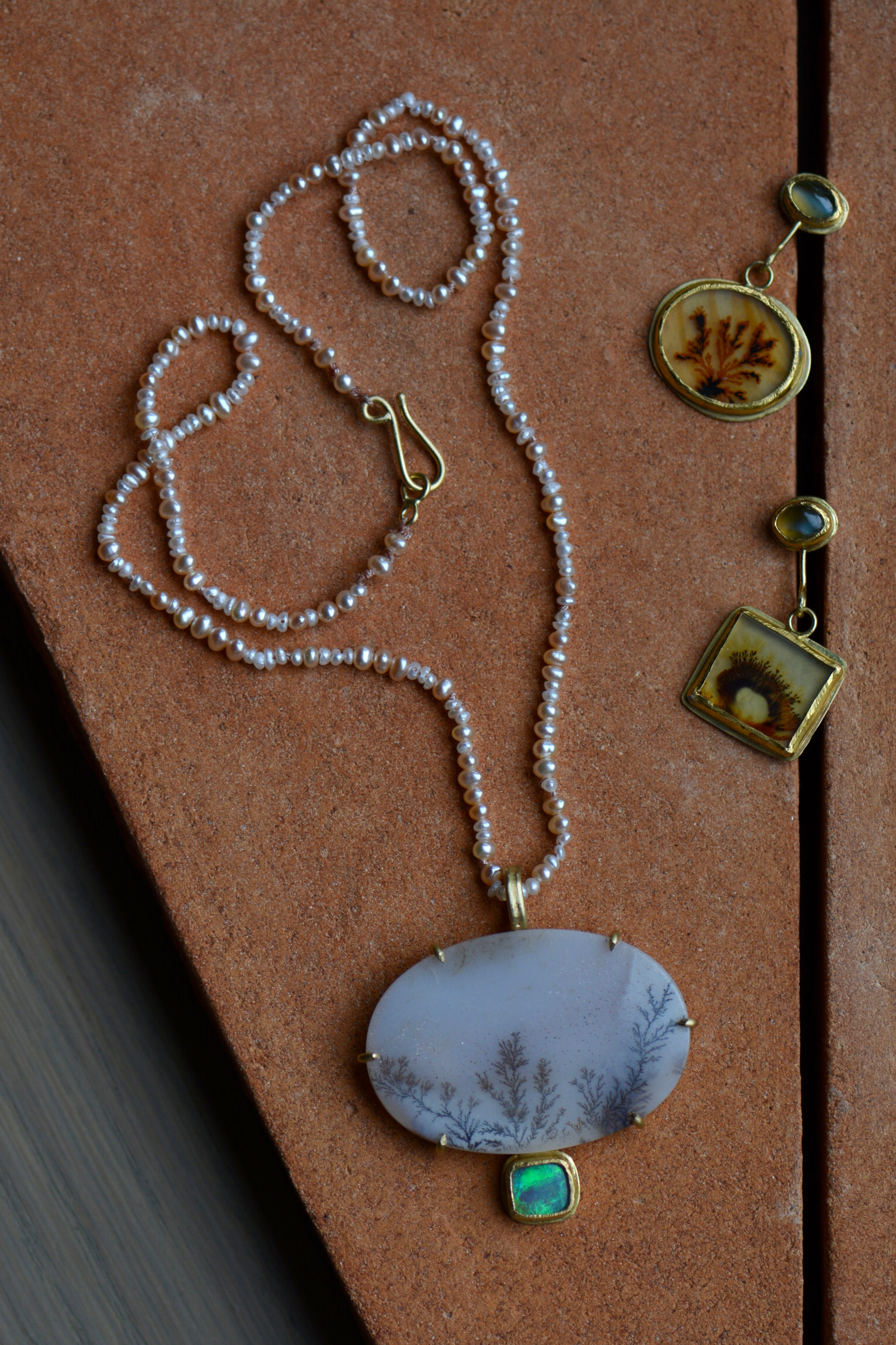 A dendritic agate and opal pendant is shown on a pearl beaded strand alongside a complimentary pair of mismatched dendritic agate and chrysoberyl earrings, all set in solid gold.