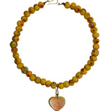 A beaded necklace made of round yellow beads with small red and green speckles, featuring a dangling heart-shaped pendant with a peach and white marbled pattern and a gold setting, shown on a plain white background.