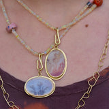 A close-up of a person wearing layered necklaces. The central necklaces have two oval gold-tone pendants with pale grey and orange dendritic agate stones, layered over a beaded opal necklace strands with pink and coral accent beads, and a gold chain necklace with B-shaped links.