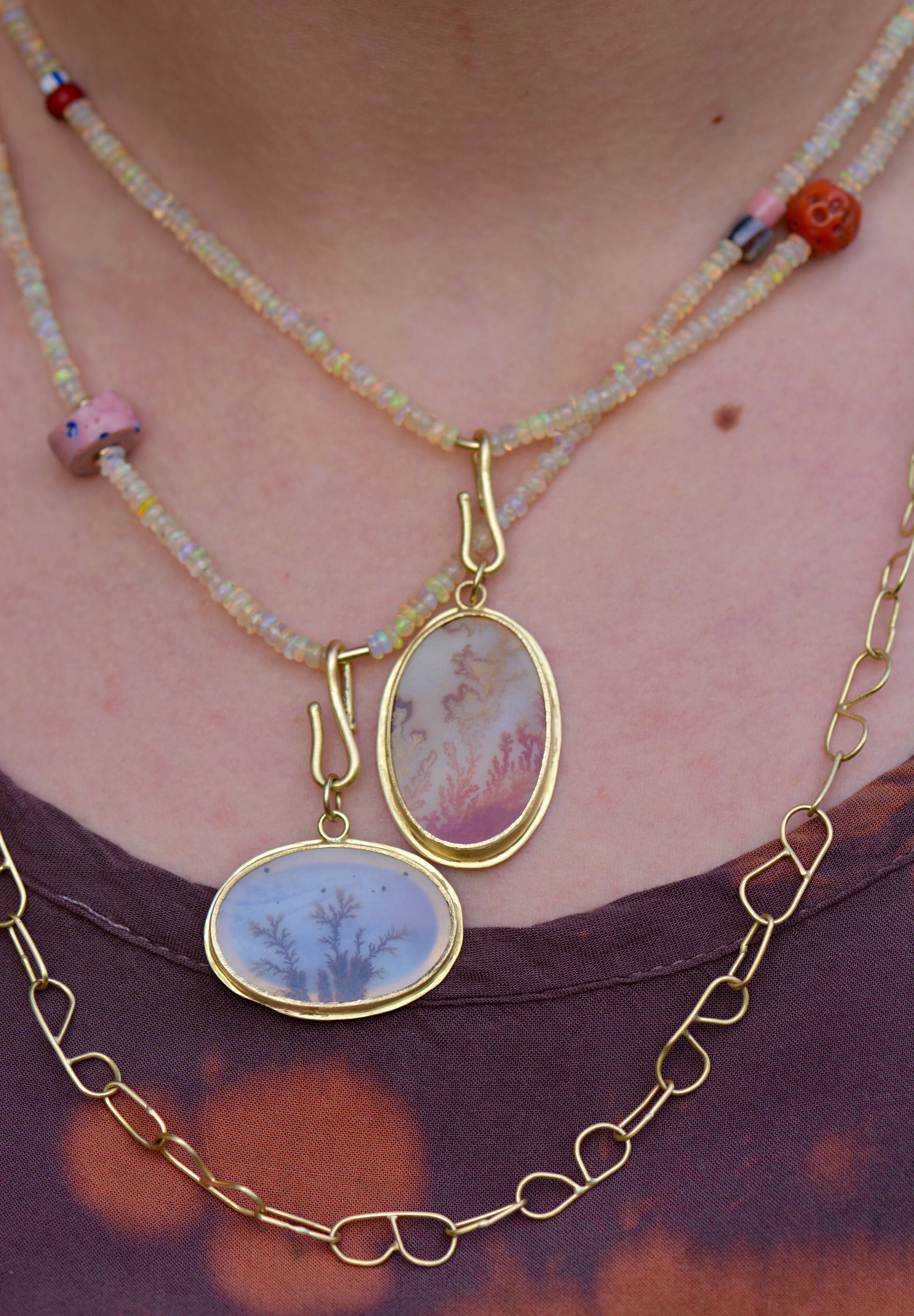 A close-up of a person wearing layered necklaces. The central necklaces have two oval gold-tone pendants with pale grey and orange dendritic agate stones, layered over a beaded opal necklace strands with pink and coral accent beads, and a gold chain necklace with B-shaped links.