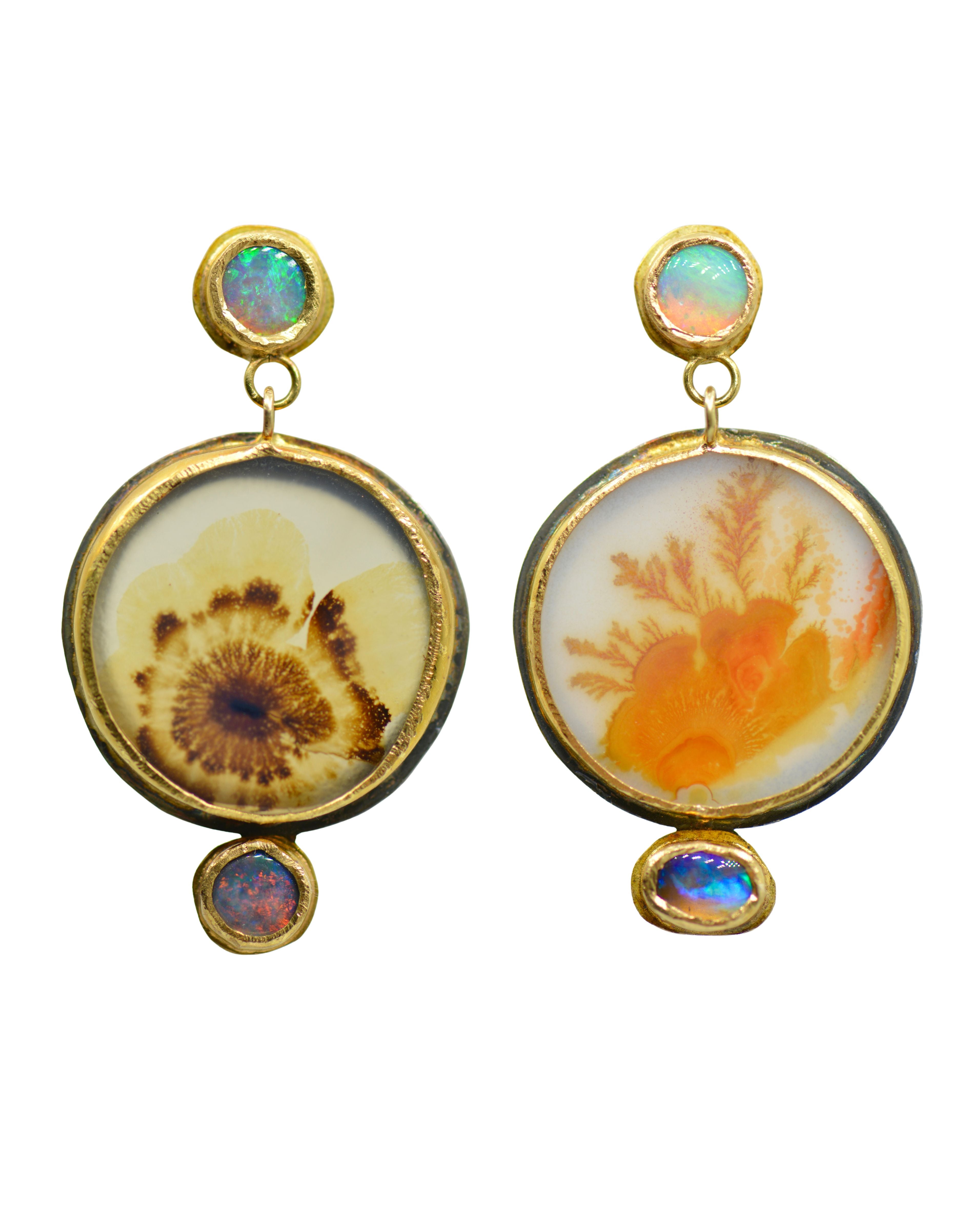 A pair of gold dangle earrings featuring dendritic agate cabochons with natural inclusions, one with a brown radiating pattern and the other with orange coral-like structures, each accented with small round iridescent opal stones at the top and bottom.