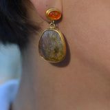 A close-up of a person's ear wearing a gold drop earring with a small round orange stone and a larger irregular beige stone pendant.