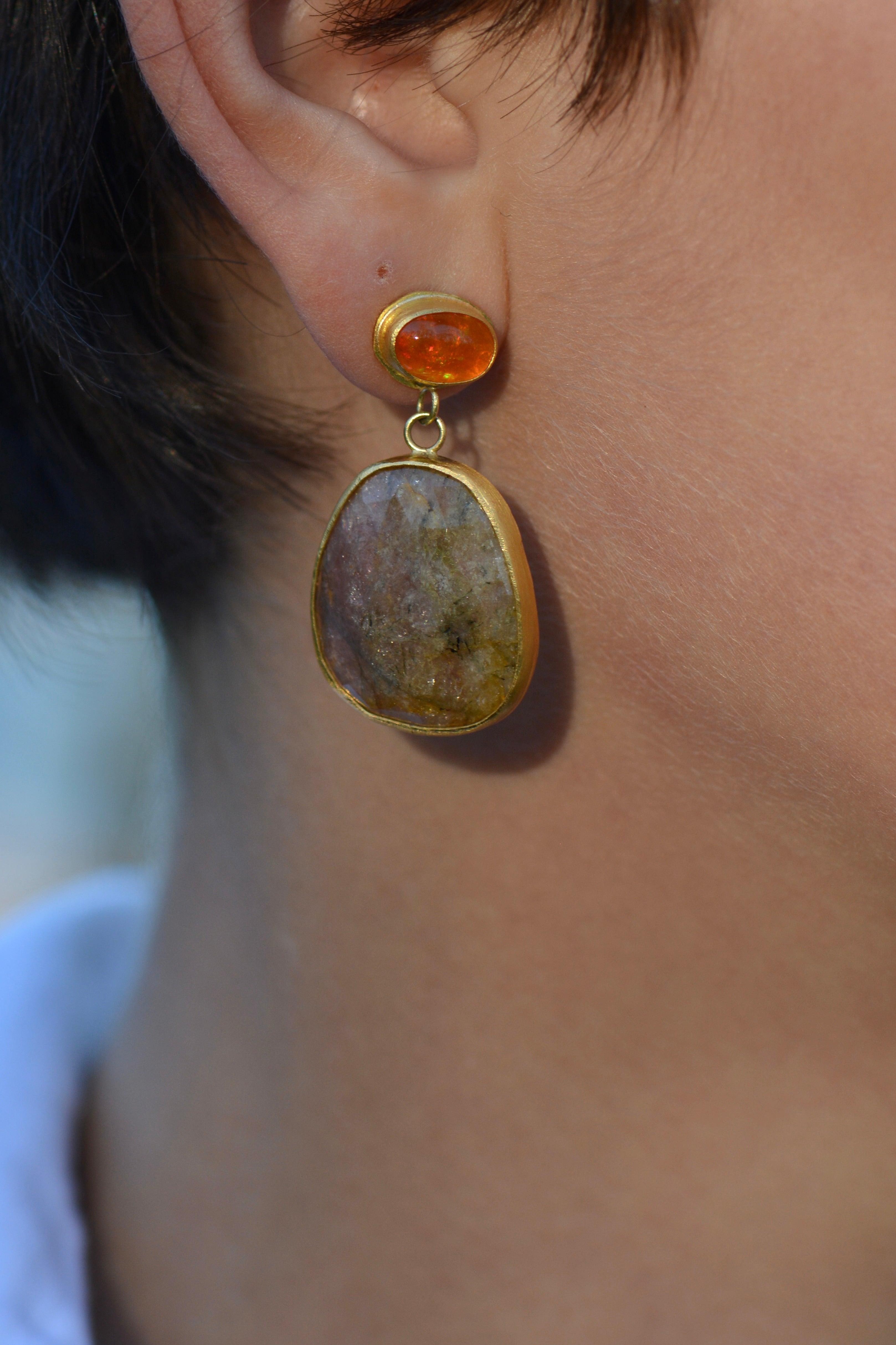 A close-up of a person's ear wearing a gold drop earring with a small round orange stone and a larger irregular beige stone pendant.