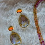Gold-tone drop earrings with oval faceted orange stones, displayed on textured cream lace next to a beaded strand necklace.