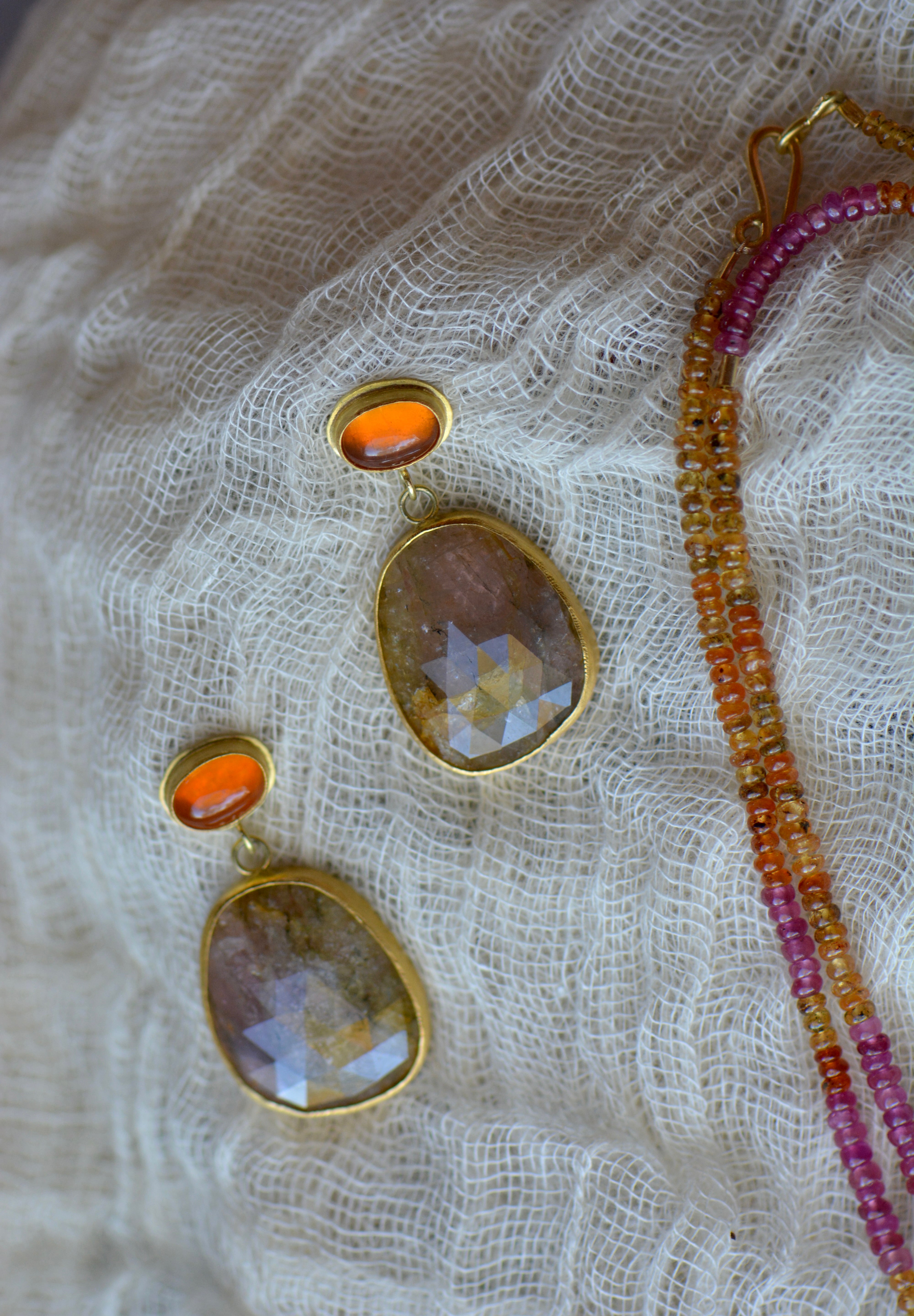 Gold-tone drop earrings with oval faceted orange stones, displayed on textured cream lace next to a beaded strand necklace.