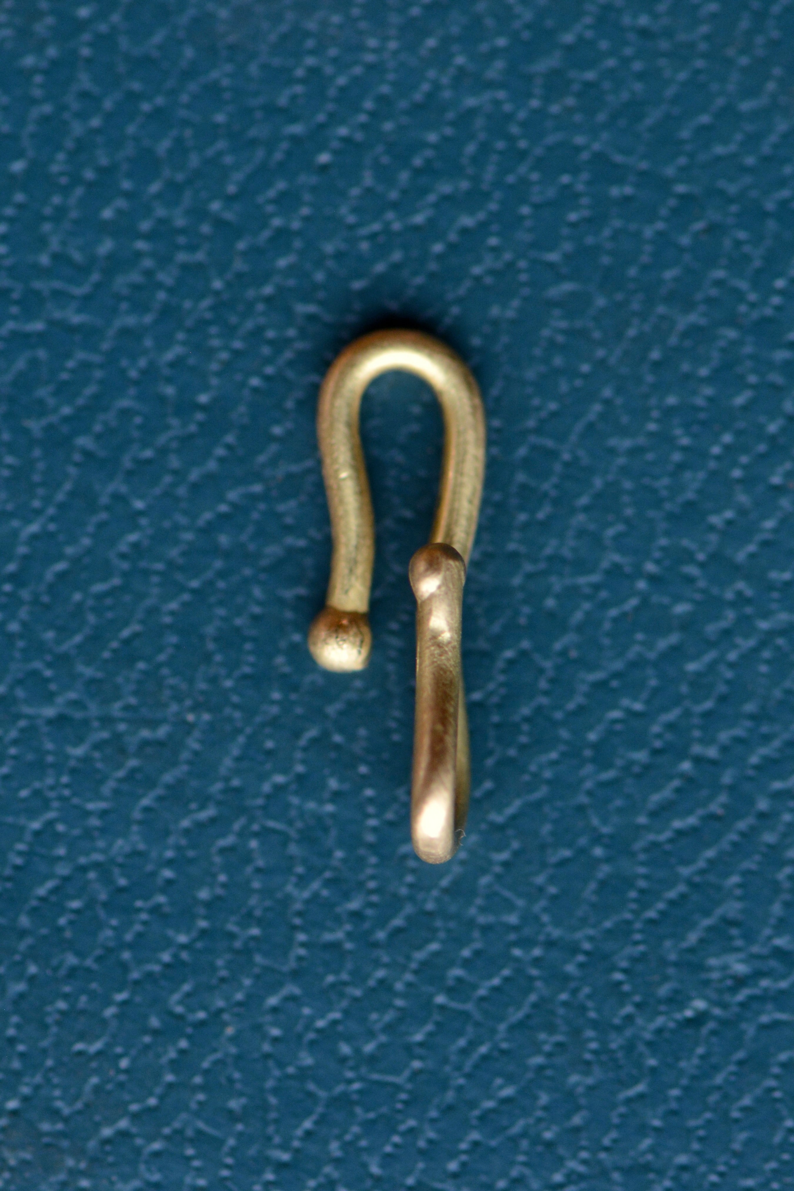 A gold charm connector hook sits on a teal blue textured backdrop.