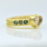 A gold ring featuring a heart-shaped clear gemstone and three small round green gemstones on each side of the heart, set against a clean white background with a soft reflection.
