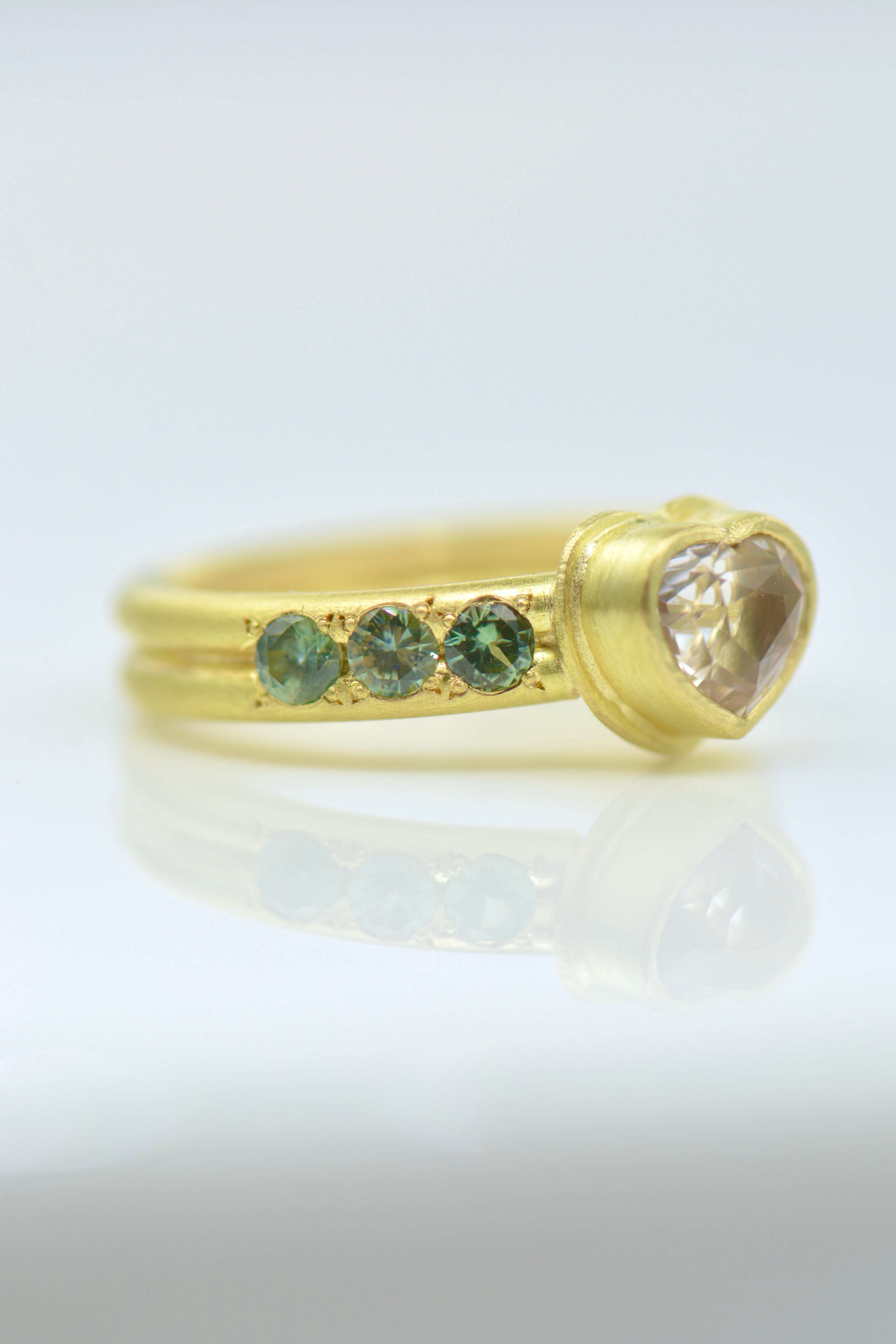 A gold ring featuring a heart-shaped clear gemstone and three small round green gemstones on each side of the heart, set against a clean white background with a soft reflection.