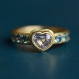 A heart-shaped centre stone in pale pink is accented with six blue round faceted stones in a gold ring setting on a teal blue backdrop.