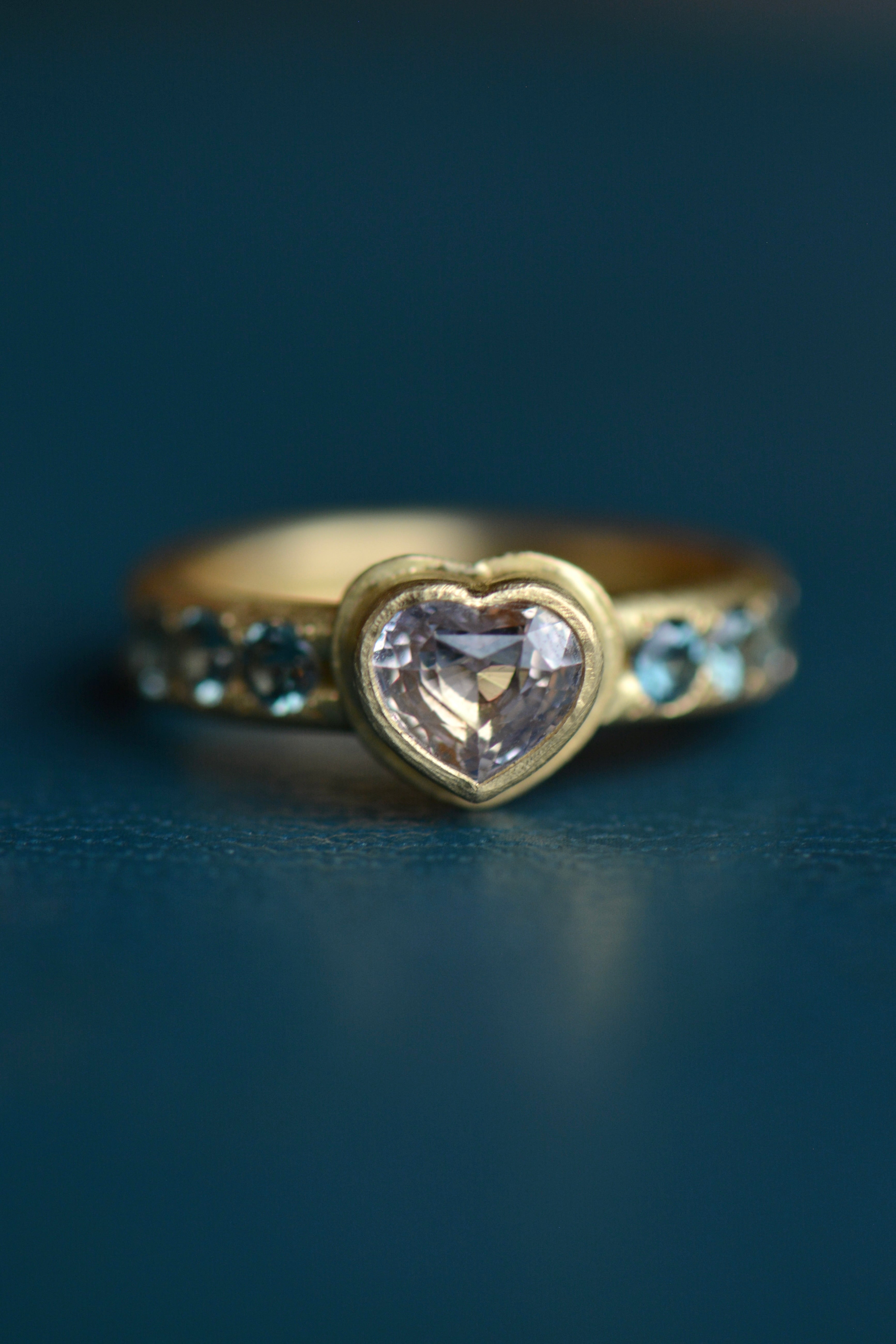 A heart-shaped centre stone in pale pink is accented with six blue round faceted stones in a gold ring setting on a teal blue backdrop.