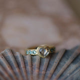 A gold ring with a heart-shaped clear pink gemstone and small blue accents, resting on a pink and grey seashell over a pinkish stone surface.