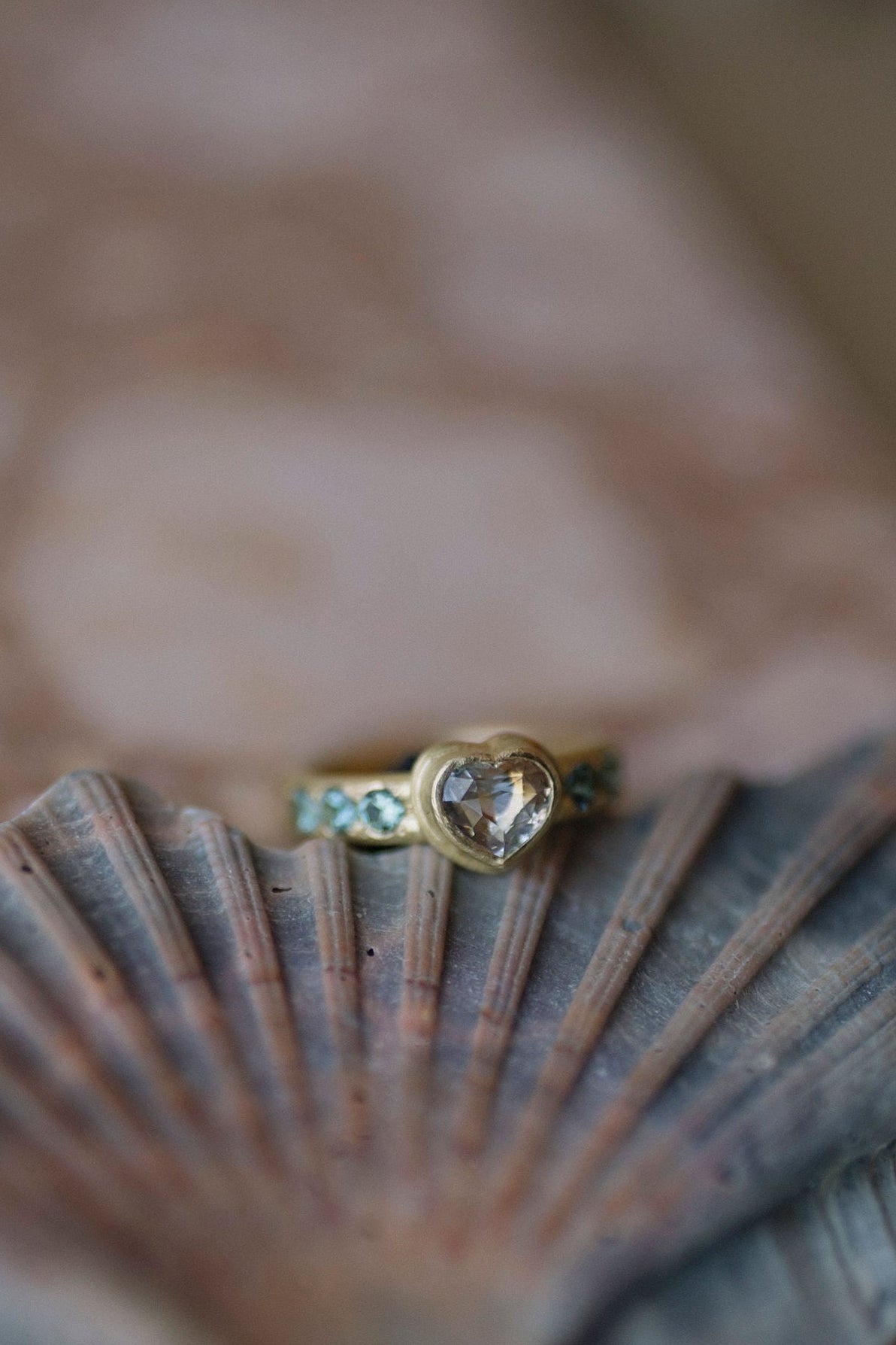A gold ring with a heart-shaped clear pink gemstone and small blue accents, resting on a pink and grey seashell over a pinkish stone surface.