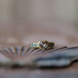A gold ring with a heart-shaped clear gemstone and teal blue accent stones, resting on a seashell against a soft, blurred background.