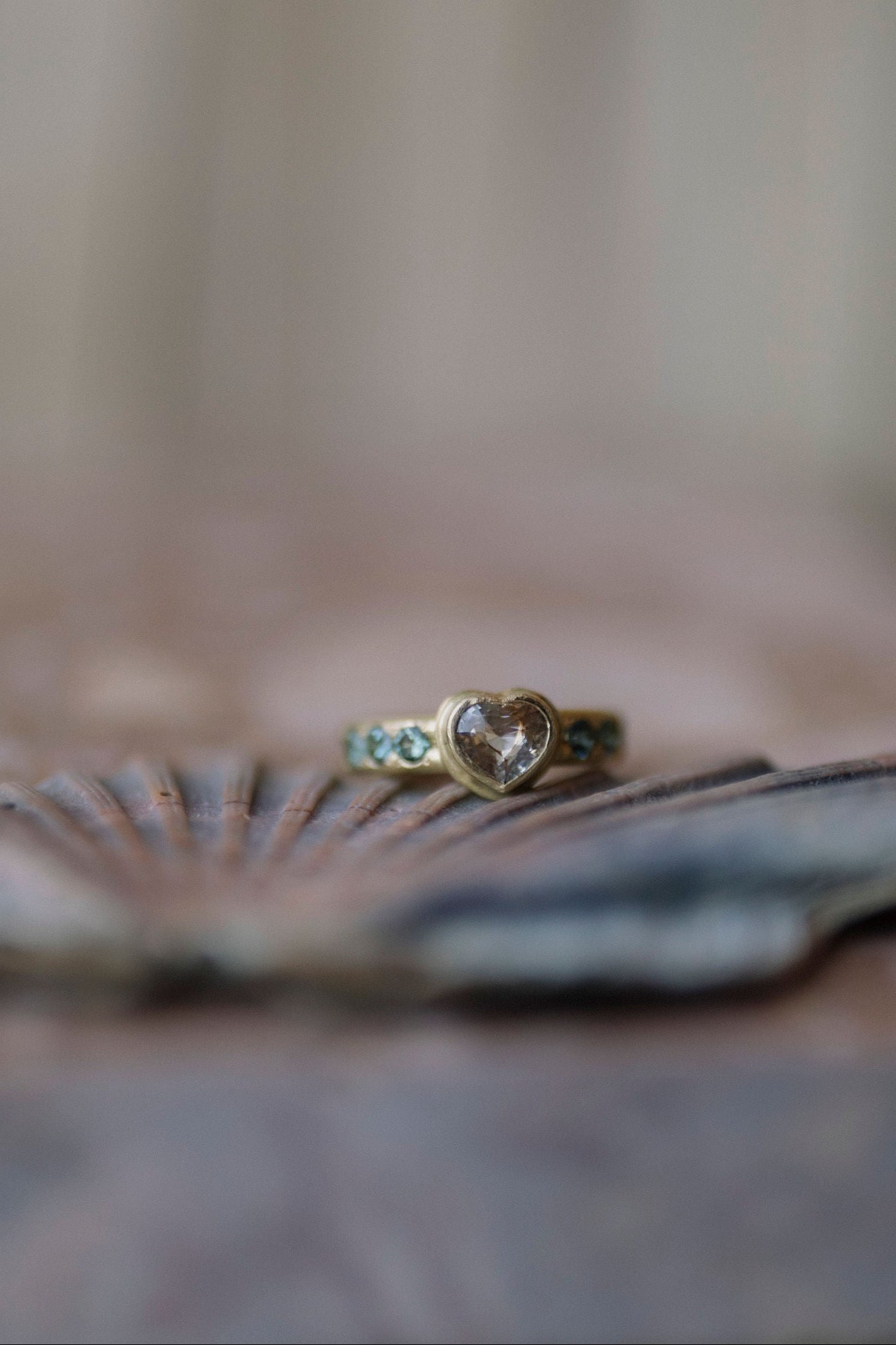 A gold ring with a heart-shaped clear gemstone and teal blue accent stones, resting on a seashell against a soft, blurred background.