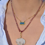 A close-up of a person wearing a layered necklace set. The necklace stack features a long, multi-strand chain with dusty rose beads and a large, irregular white pendant with a small green stone accent, layered over a thinner gold chain with a rectangular blue stone pendant.