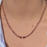 A close-up of a person's neck and upper chest wearing a delicate beaded necklace with small purplish-brown beads and a central gold accent bead, paired with a white top.