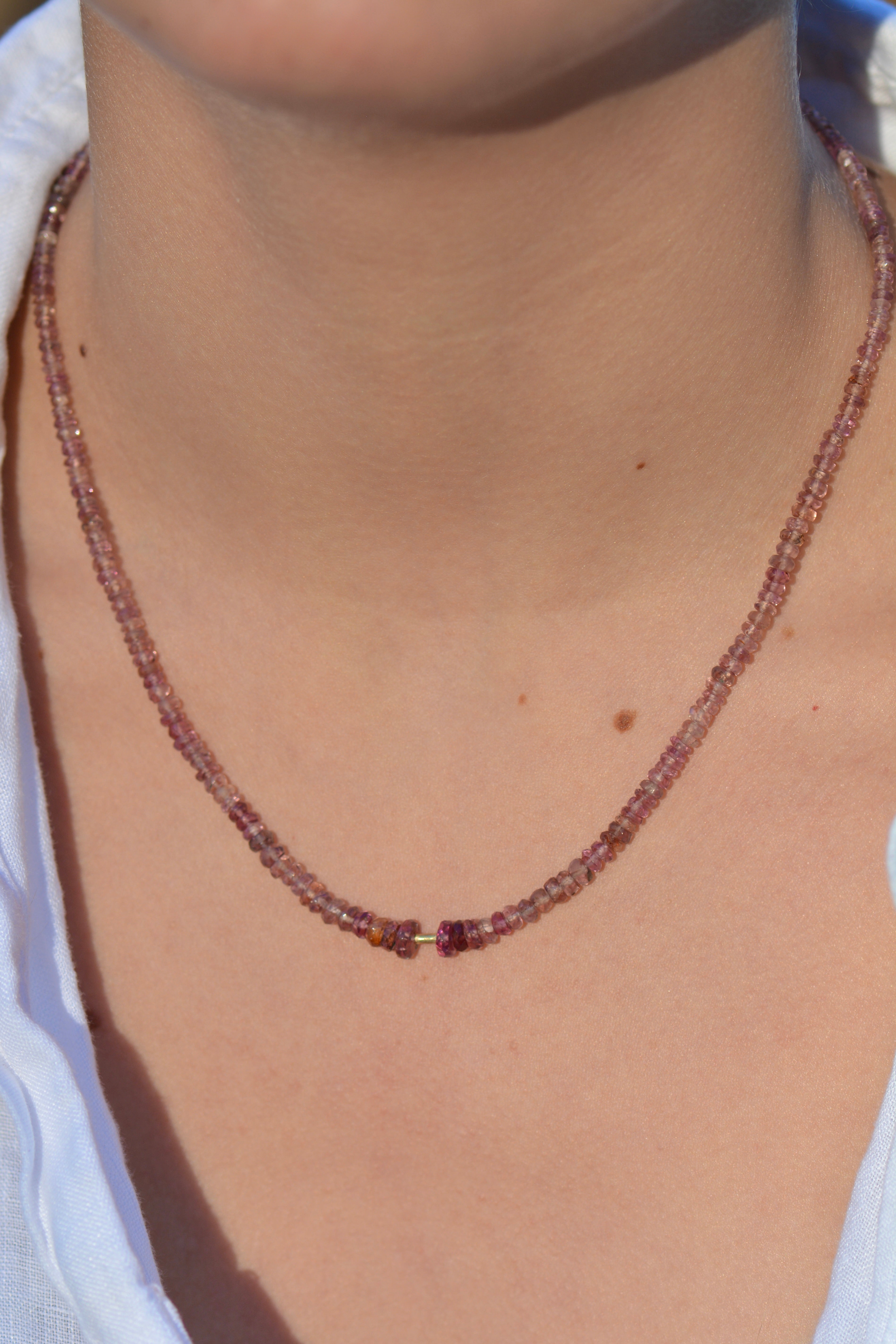 A close-up of a person's neck and upper chest wearing a delicate beaded necklace with small purplish-brown beads and a central gold accent bead, paired with a white top.