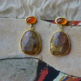 A pair of gold drop earrings with oval orange top stones and larger freeform-shaped brown, pink and gold marbled stone pendants, displayed on a textured beige and red surface.