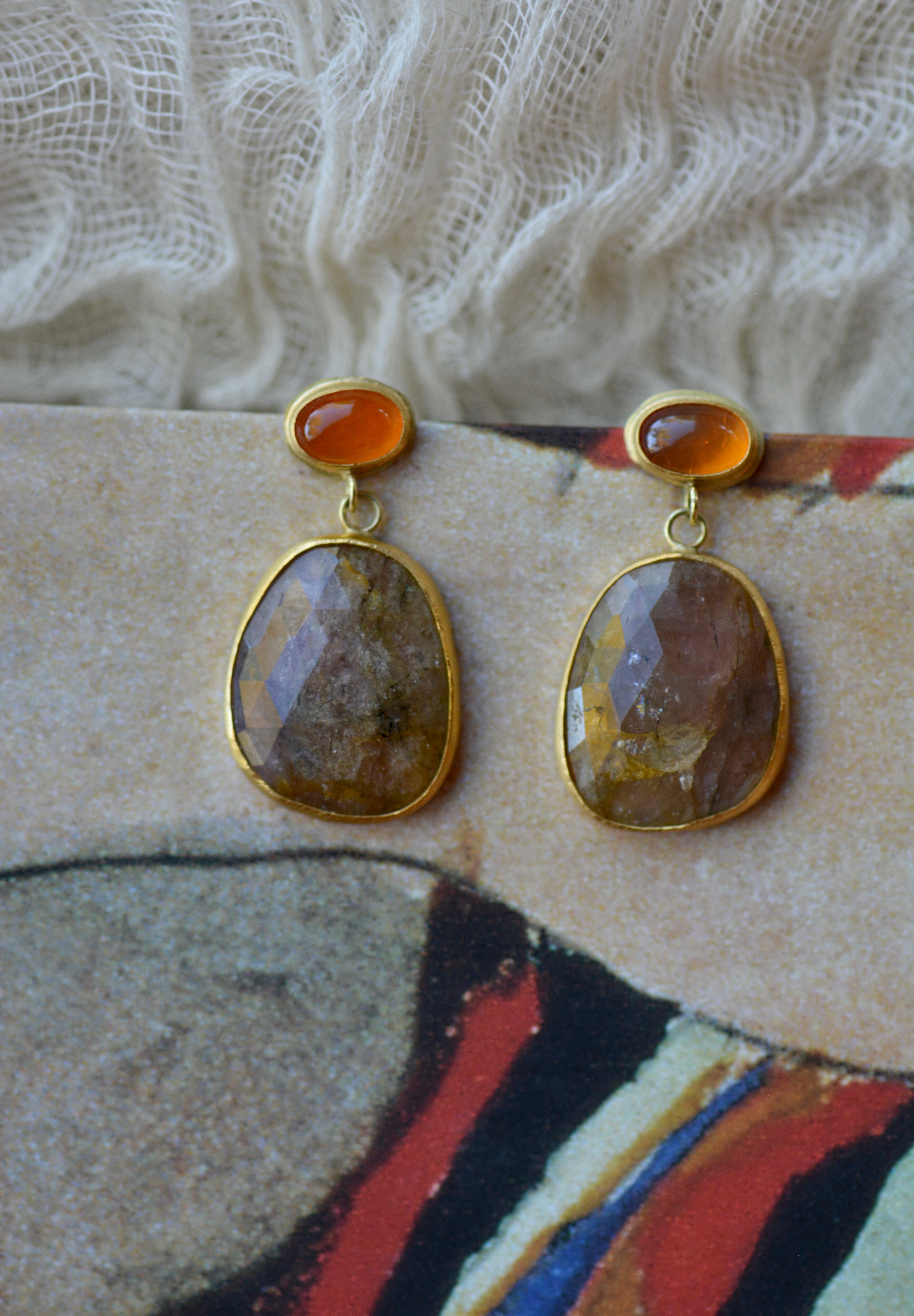 A pair of gold drop earrings with oval orange top stones and larger freeform-shaped brown, pink and gold marbled stone pendants, displayed on a textured beige and red surface.