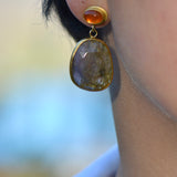 A close-up of a person's ear wearing a gold drop earring with a small orange top cabochon and a larger irregular brownish-pink stone below with light glowing through.