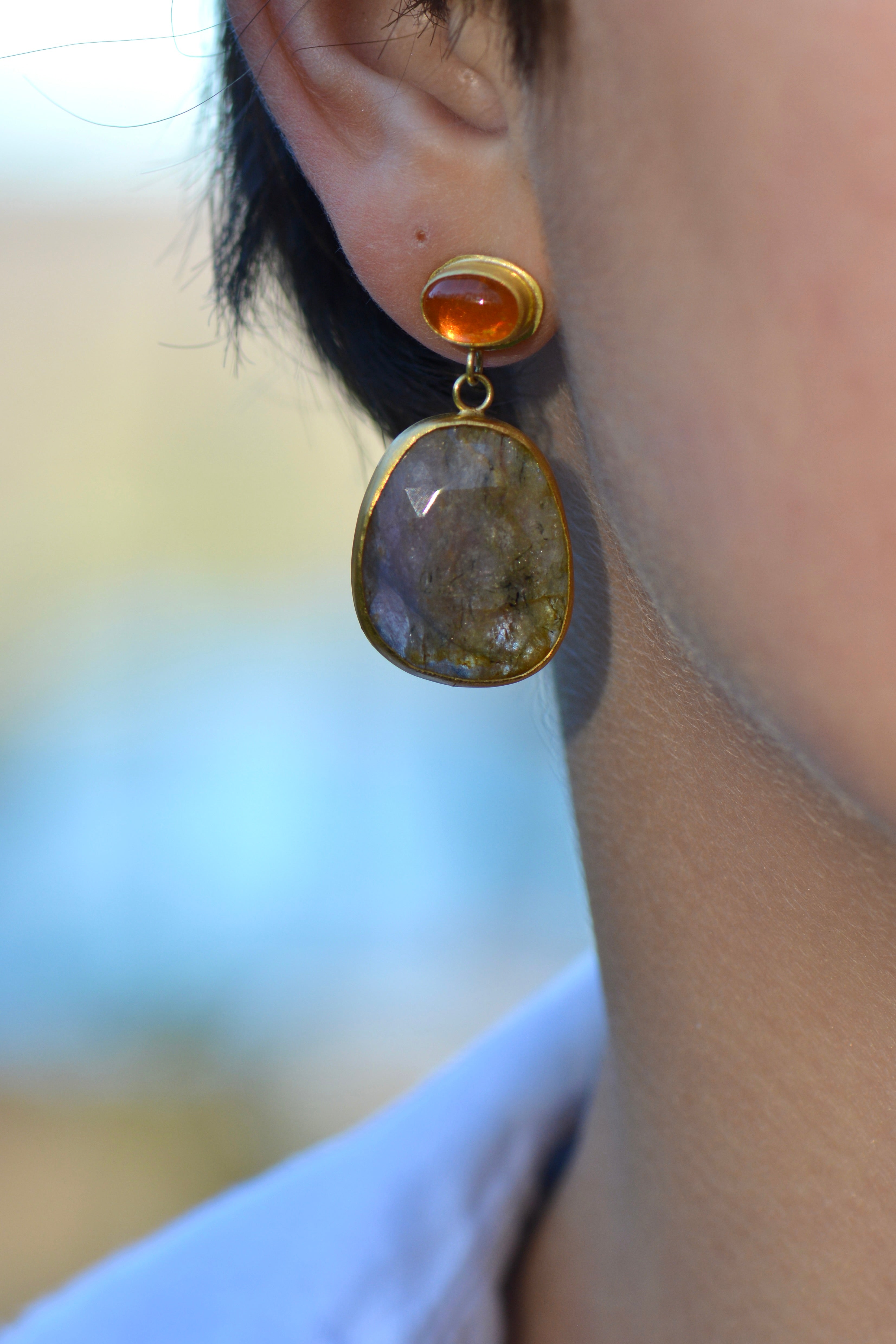 A close-up of a person's ear wearing a gold drop earring with a small orange top cabochon and a larger irregular brownish-pink stone below with light glowing through.
