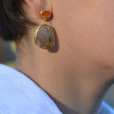A close-up of a person's ear wearing a gold drop earring with a small oval orange stone and a larger irregular beige stone pendant.