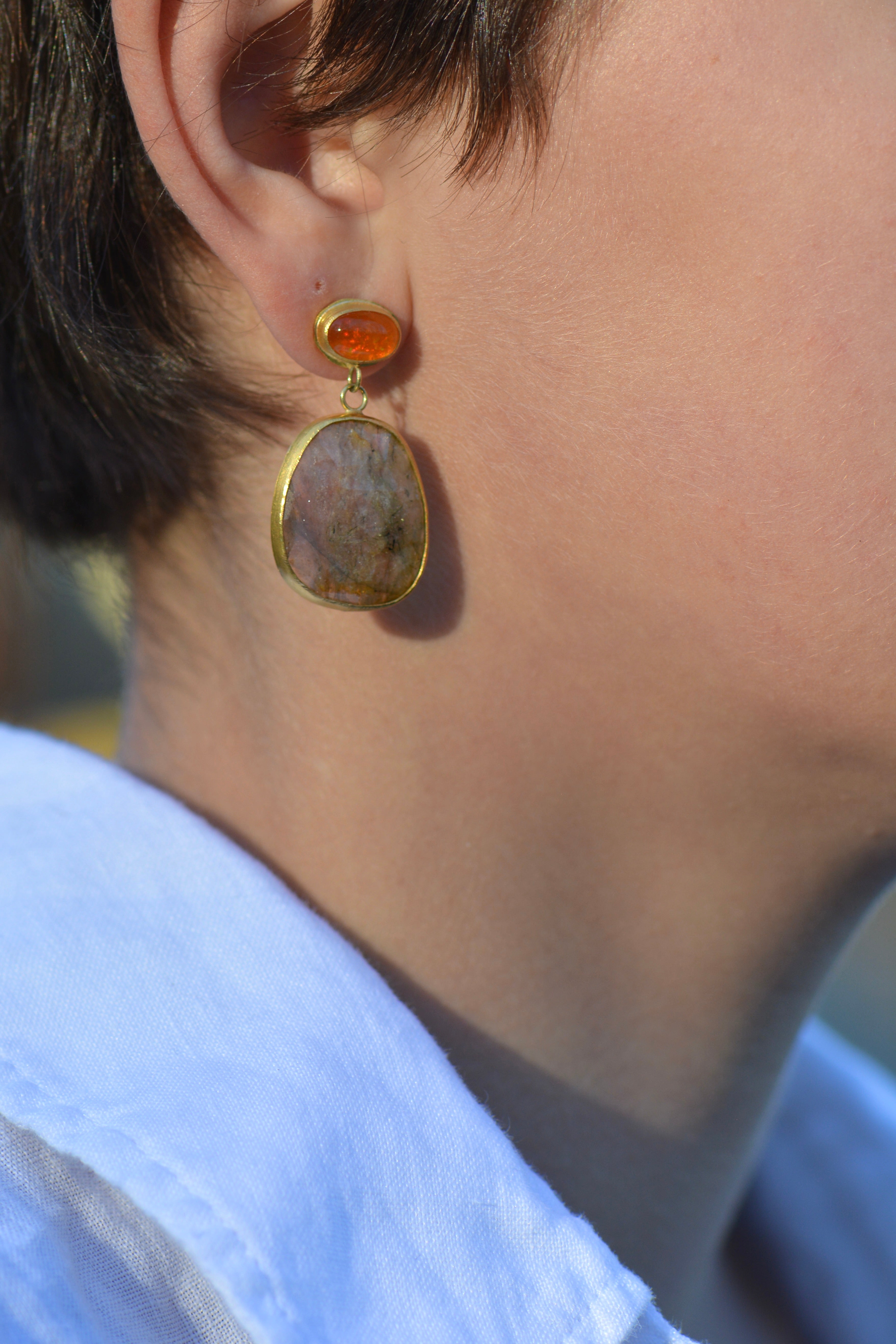 A close-up of a person's ear wearing a gold drop earring with a small oval orange stone and a larger irregular beige stone pendant.