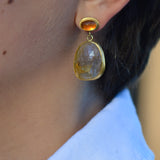 A close-up of a person's ear wearing a gold drop earring with a faceted orange top stone and a larger teardrop-shaped pink and brown stone below, paired with a white linen collared shirt.