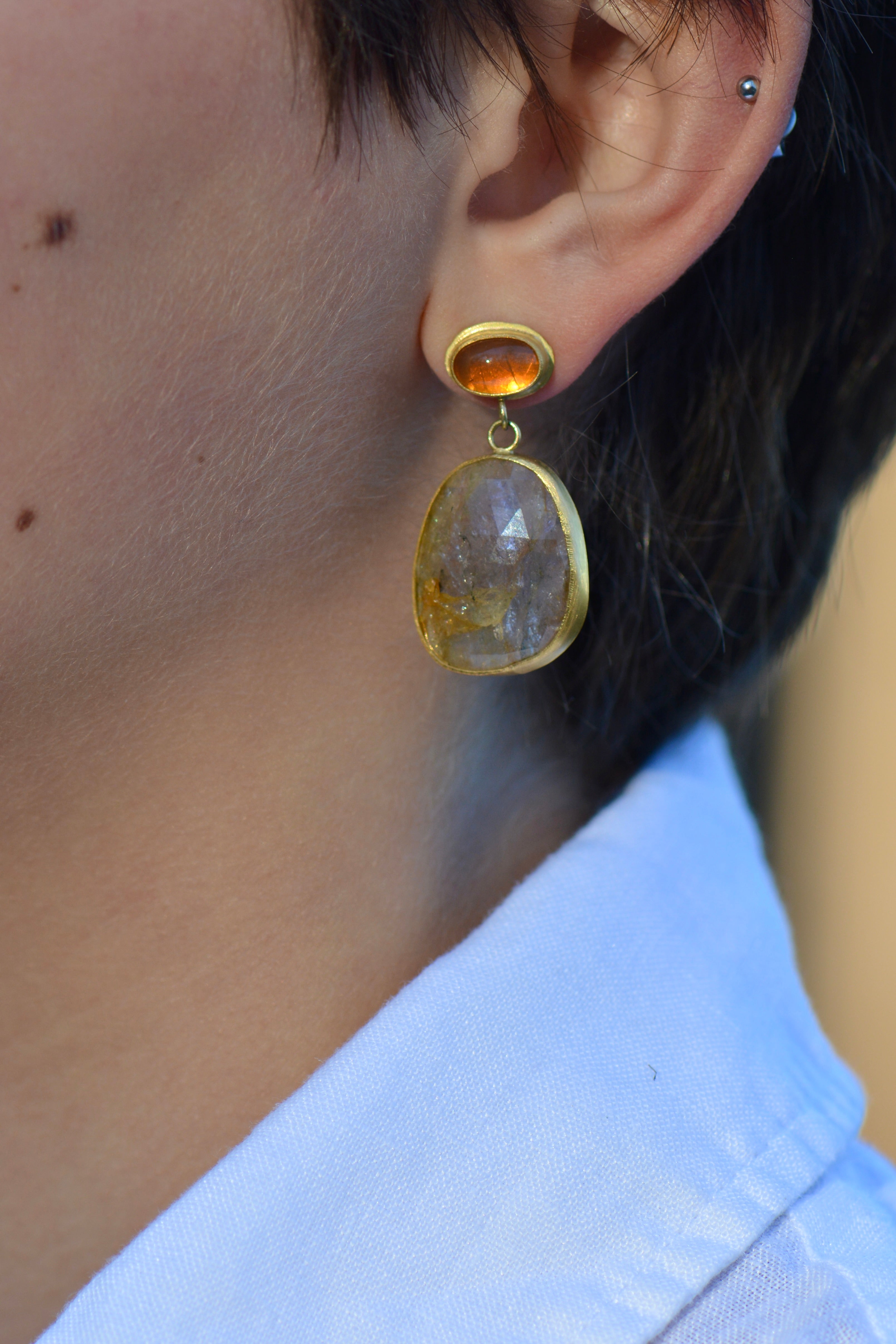A close-up of a person's ear wearing a gold drop earring with a faceted orange top stone and a larger teardrop-shaped pink and brown stone below, paired with a white linen collared shirt.