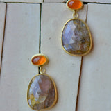 A pair of gold dangle earrings with faceted earthy pink and golden stones and bright orange cabochon accents, displayed on a light beige tiled surface.