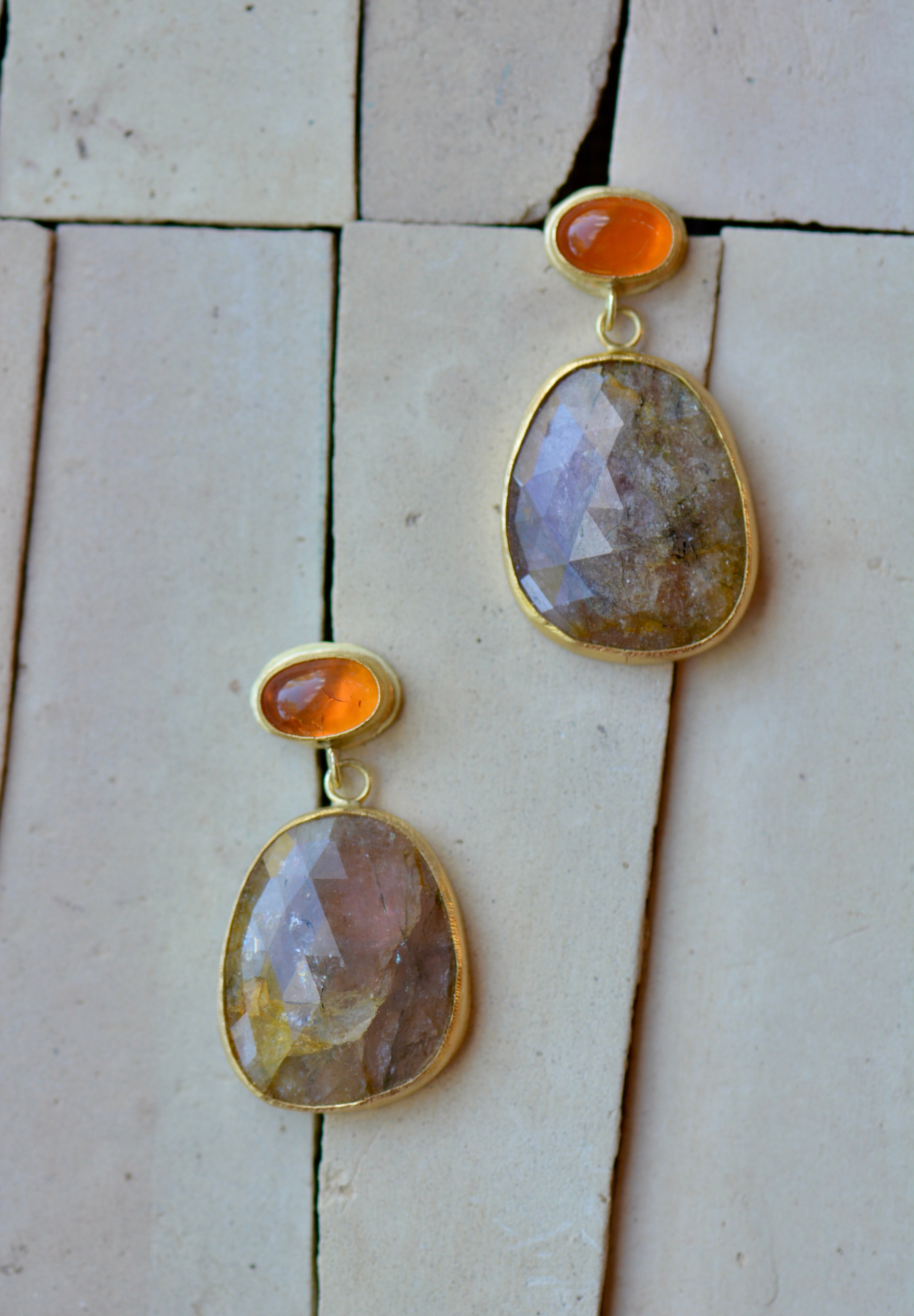 A pair of gold dangle earrings with faceted earthy pink and golden stones and bright orange cabochon accents, displayed on a light beige tiled surface.