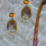 Gold-toned drop earrings with oval faceted orange stones and a beaded strand of orange and pink beads resting on textured cream gauze.