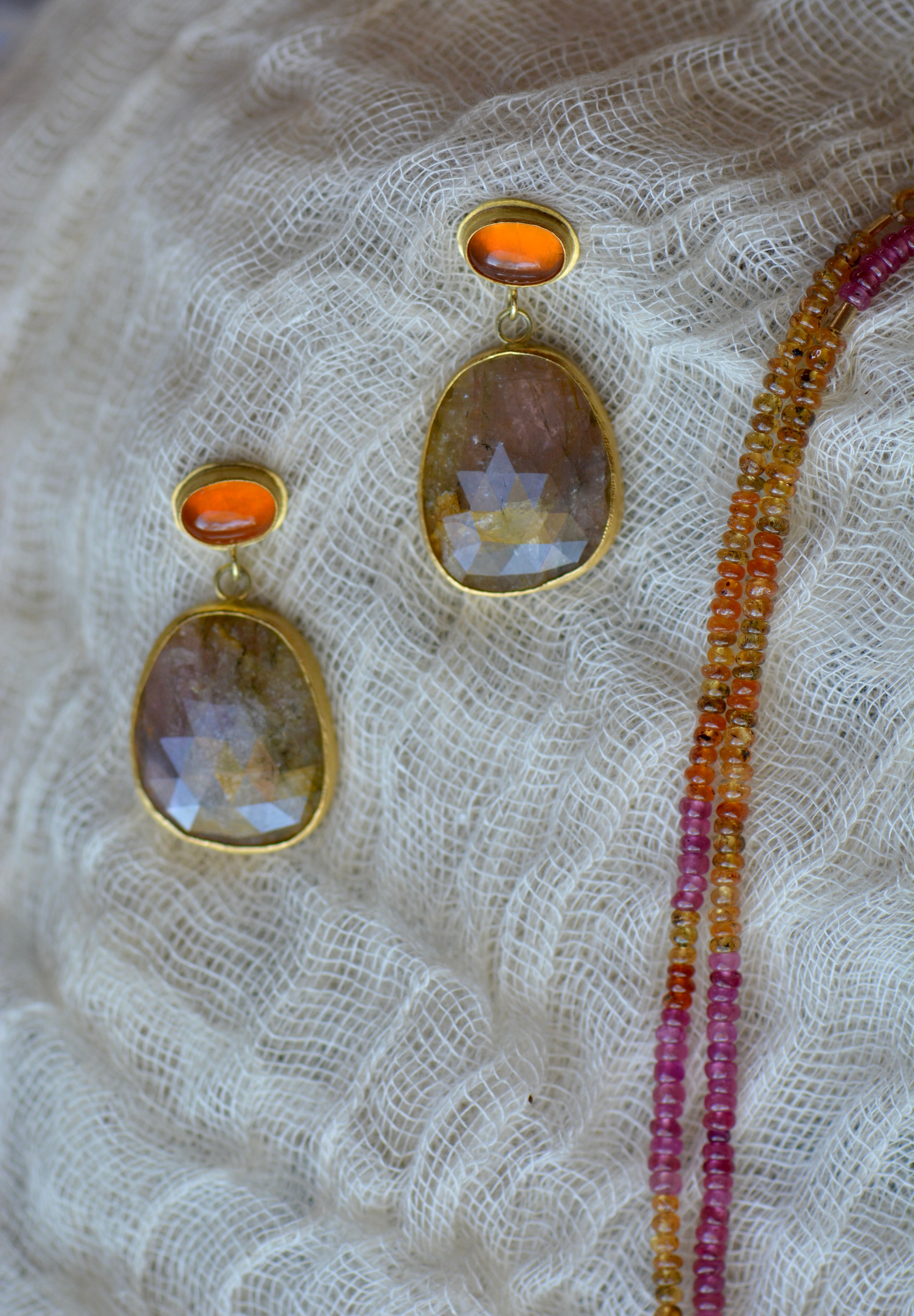 Gold-toned drop earrings with oval faceted orange stones and a beaded strand of orange and pink beads resting on textured cream gauze.
