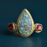 A teardrop shaped central cabochon stone in a pale turquoise colour is flanked by red faceted stones in a solid gold ring setting.