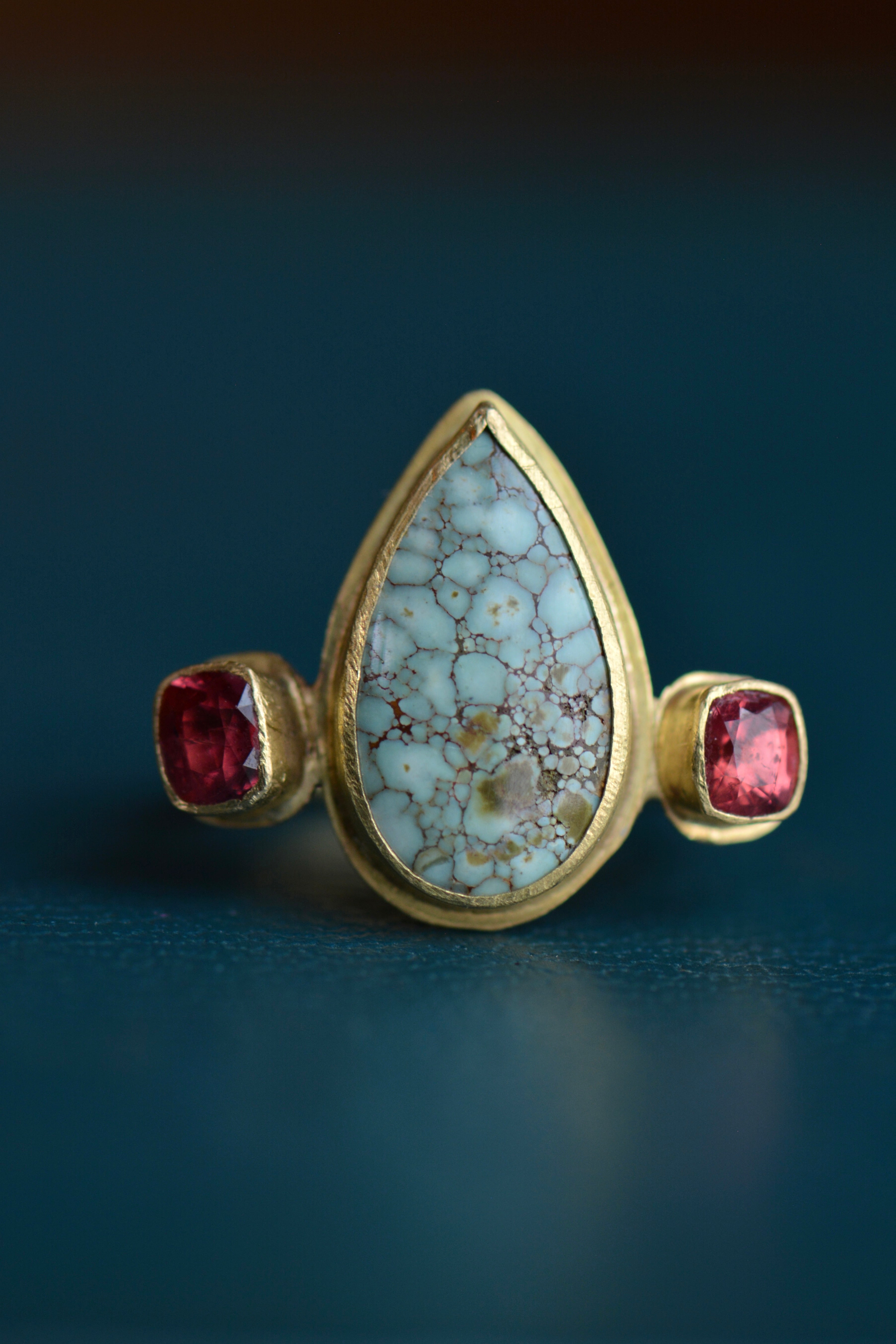 A teardrop shaped central cabochon stone in a pale turquoise colour is flanked by red faceted stones in a solid gold ring setting.