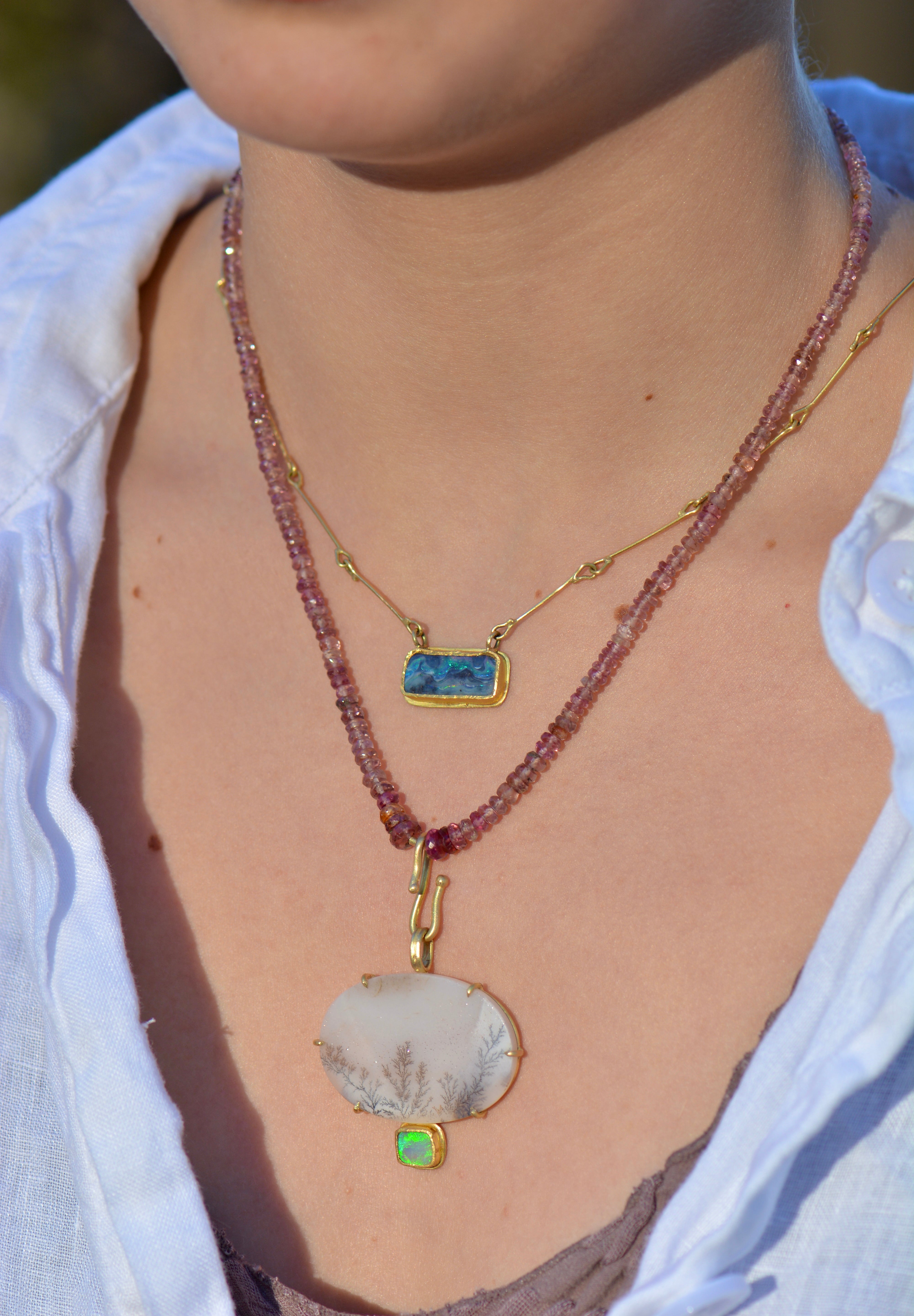 A close-up of a person wearing a layered necklace set. The set features a long, dusty rose bead strand with a large, irregular white agate pendant with a small green stone accent, layered over a thinner gold chain with a rectangular blue stone pendant.