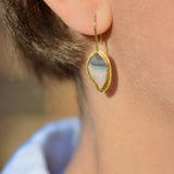 A close-up of a person's ear wearing a gold drop earring with a teardrop-shaped iridescent stone that shifts between pale blue, pink, and white.