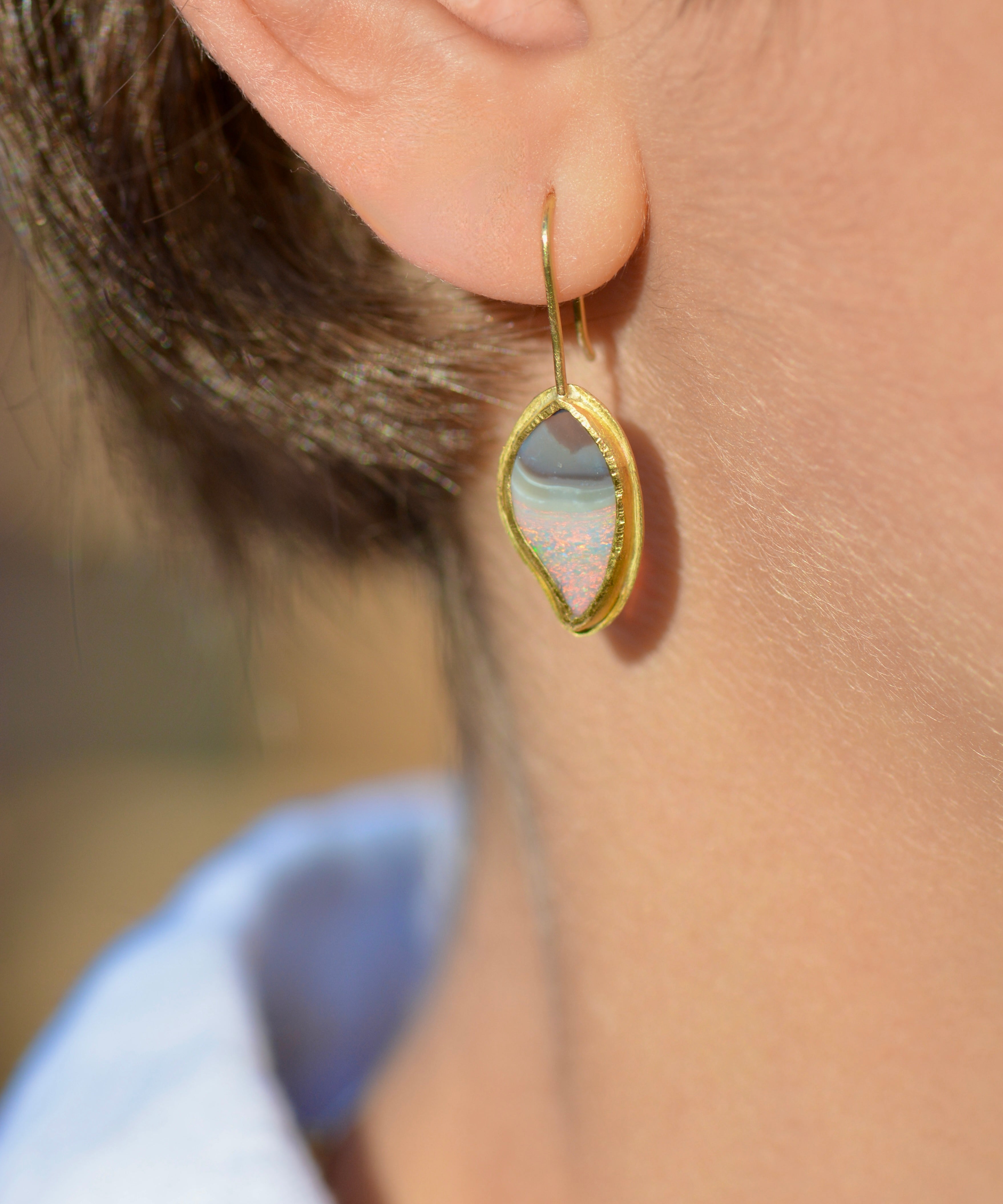 A close-up of a person's ear wearing a gold drop earring with a teardrop-shaped iridescent stone that shifts between pale blue, pink, and white.
