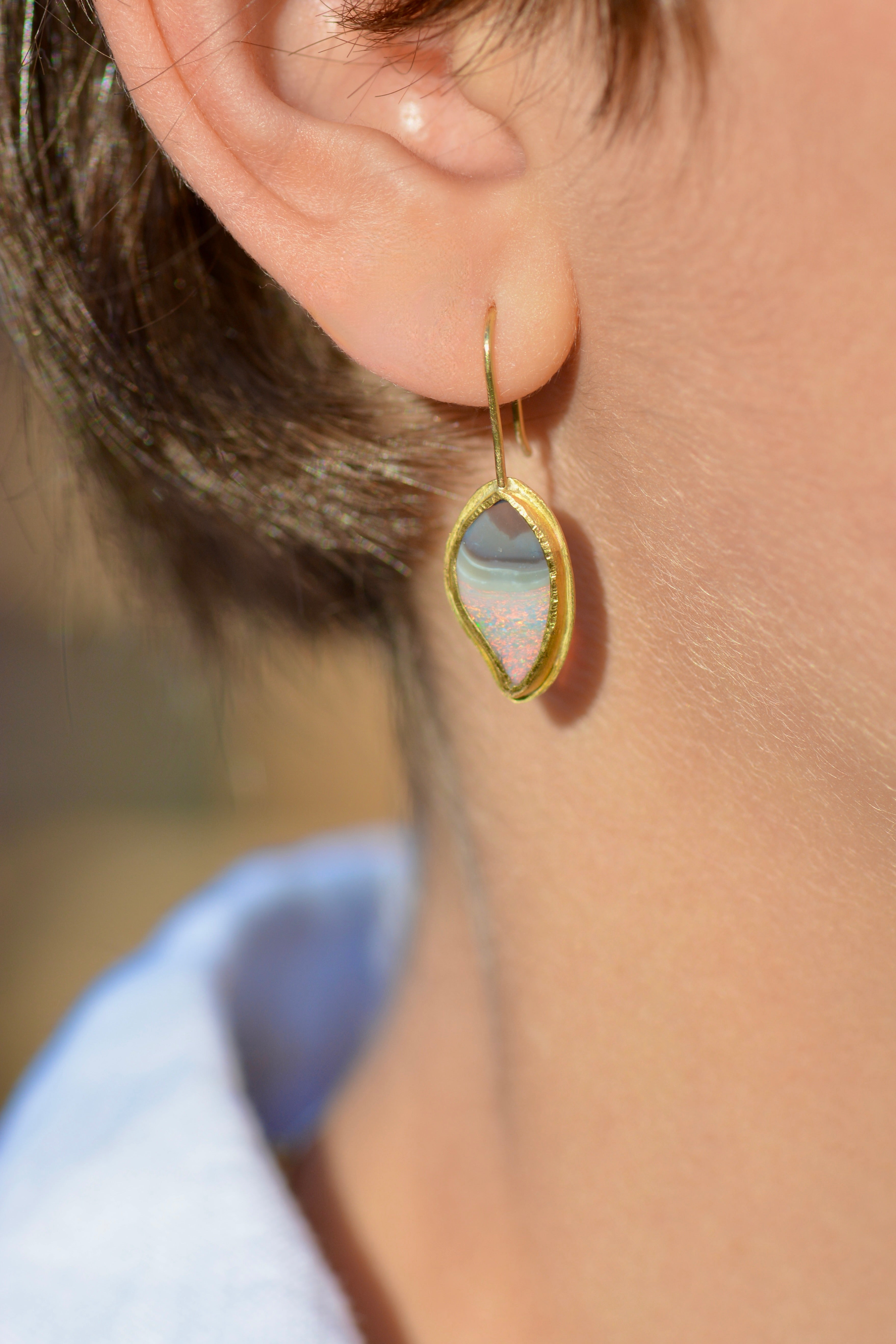 A close-up of a person's ear wearing a gold drop earring with a teardrop-shaped iridescent stone that shifts between pale blue, pink, and white.